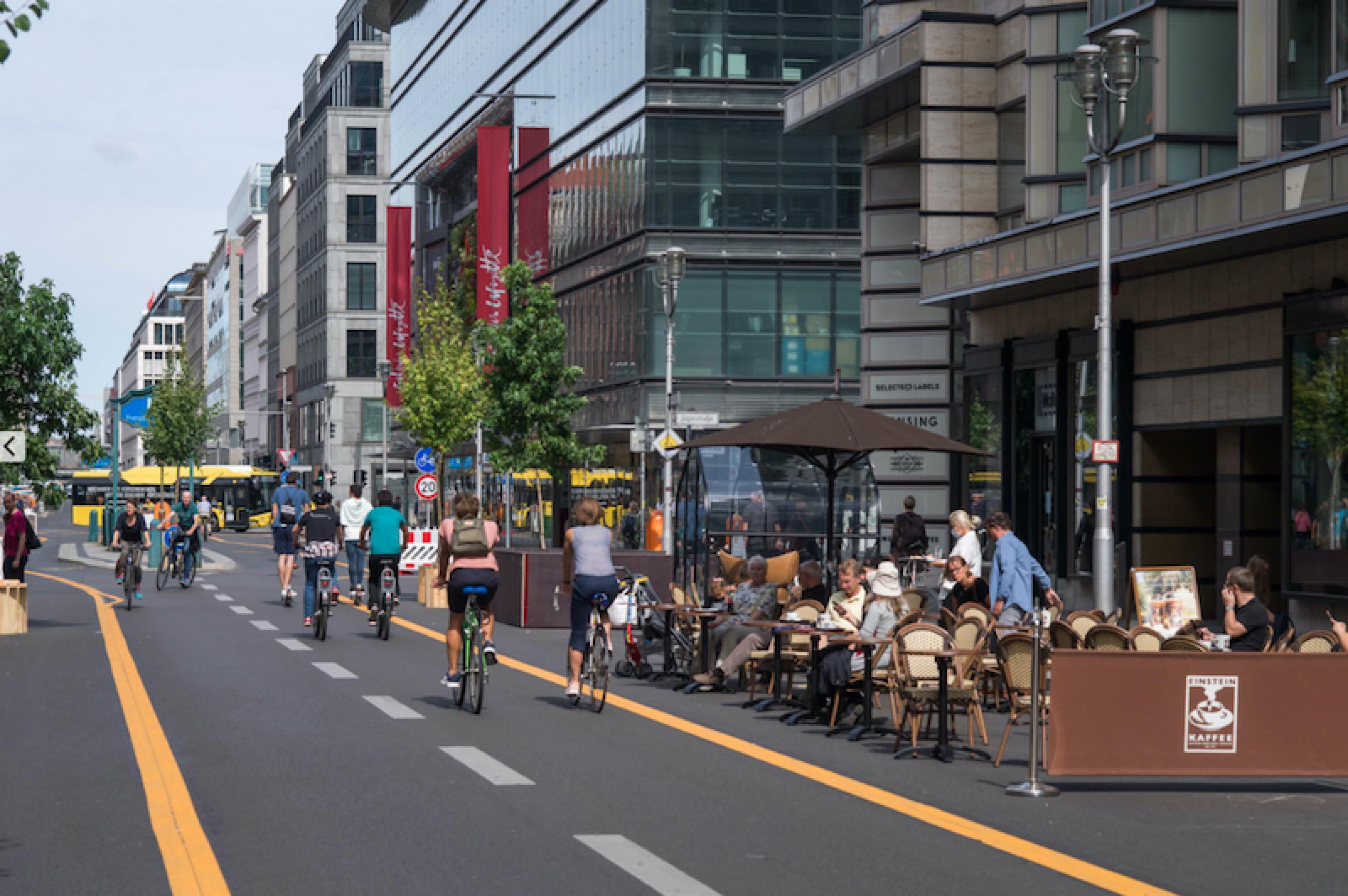 Menschen sitzen in Straßencafés in der Sonne und Radfahrer fahren auf der Fahrbahn an ihnen vorbei