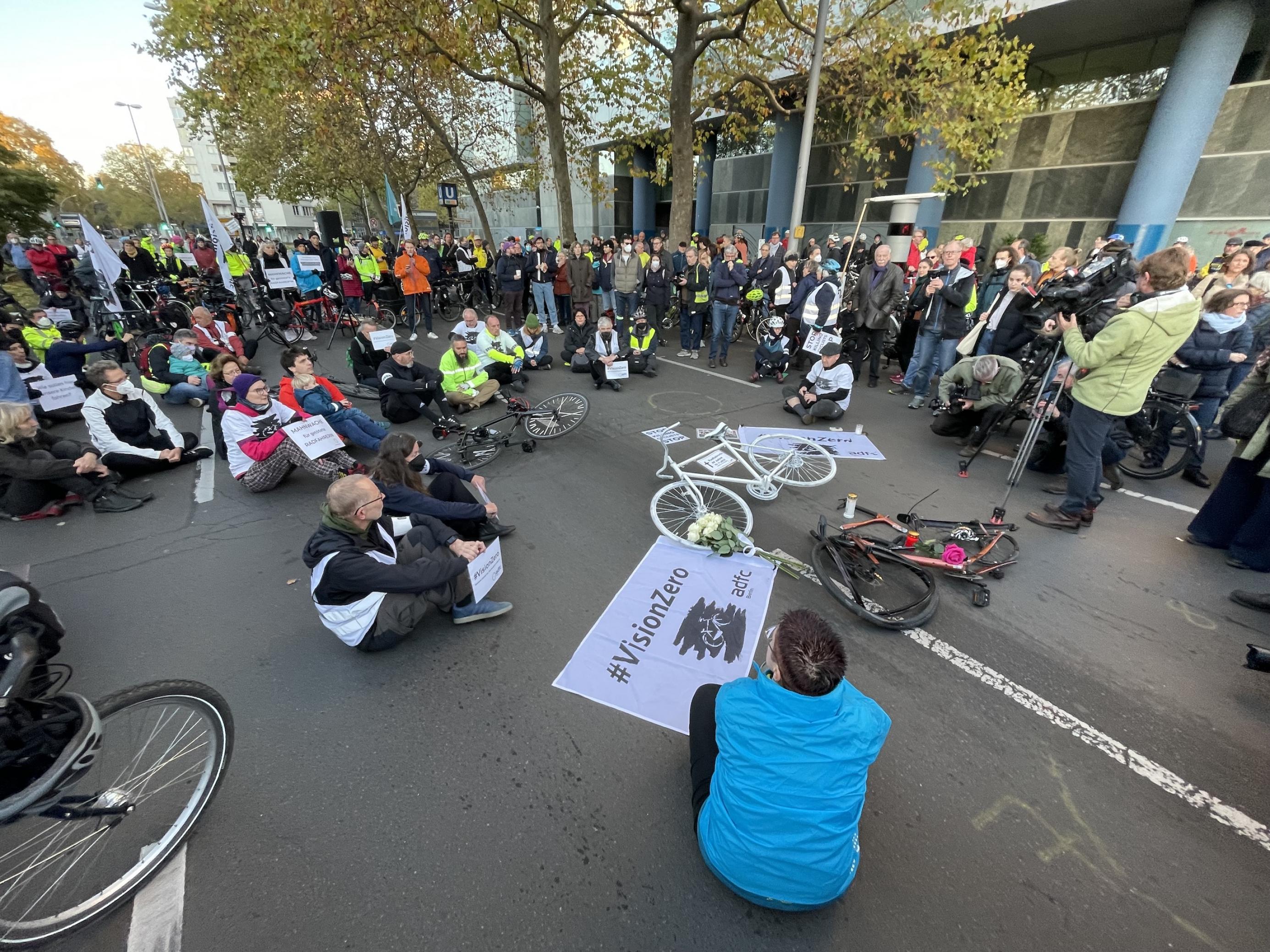 Eine Menschenmange versammelt sich um ein komplett weiß bemaltes Fahrrad, das mitten auf der Straße liegt. Daneben ein Banner #VisionZero für das Ziel, dass es Null Verkehrstote gibt.