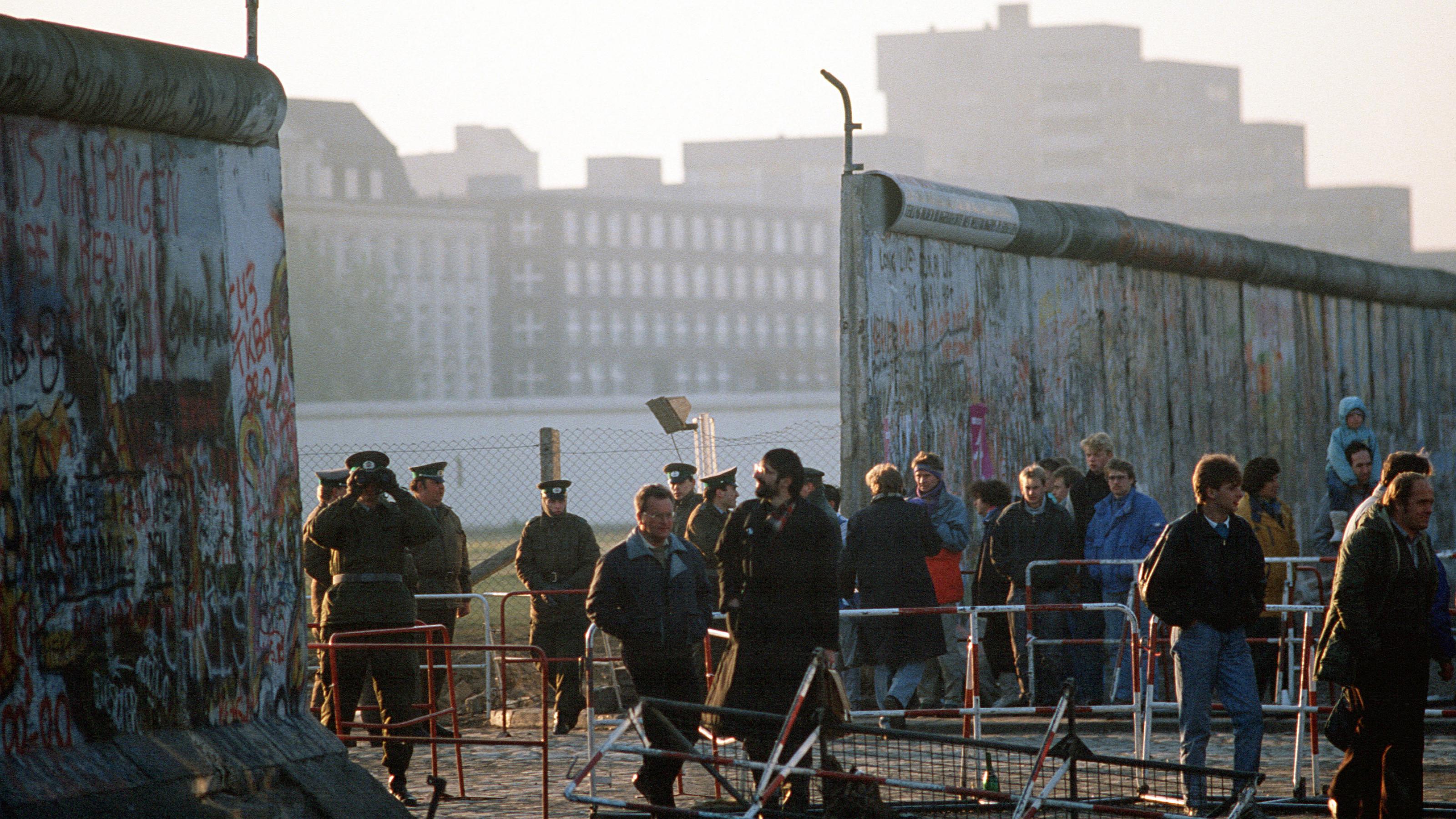 Nach dem Mauerfall gehen Bürger durch die neue Öffnung in der Berliner Mauer am Potsdamer Platz. Neben der Mauer stehen DDR-Polizisten, die die Szenerie beobachten.