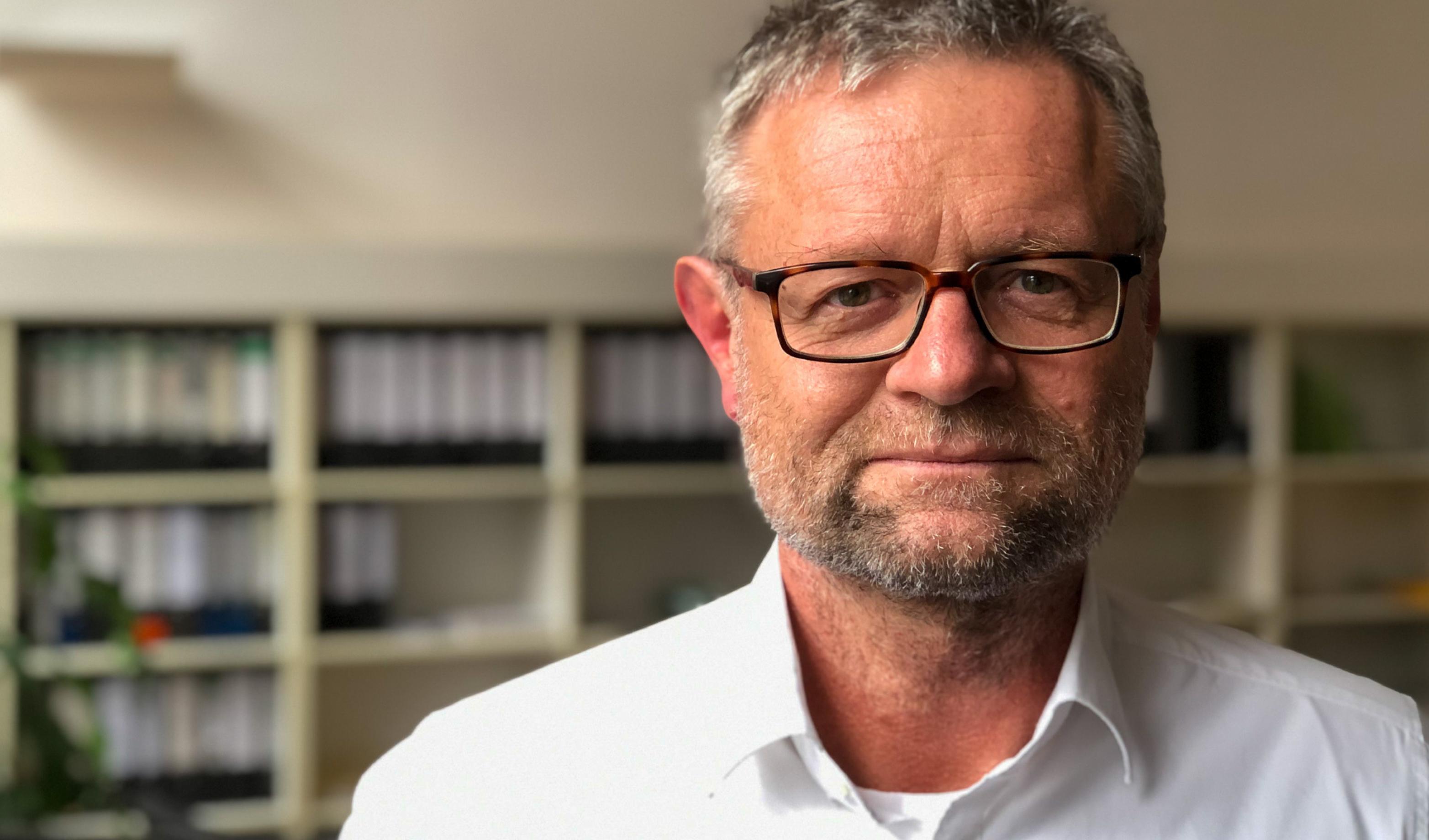 Ein Mann mit grauen Haaren und Vollbart, eckiger Brille und weißem Hemd schaut in die Kamera.