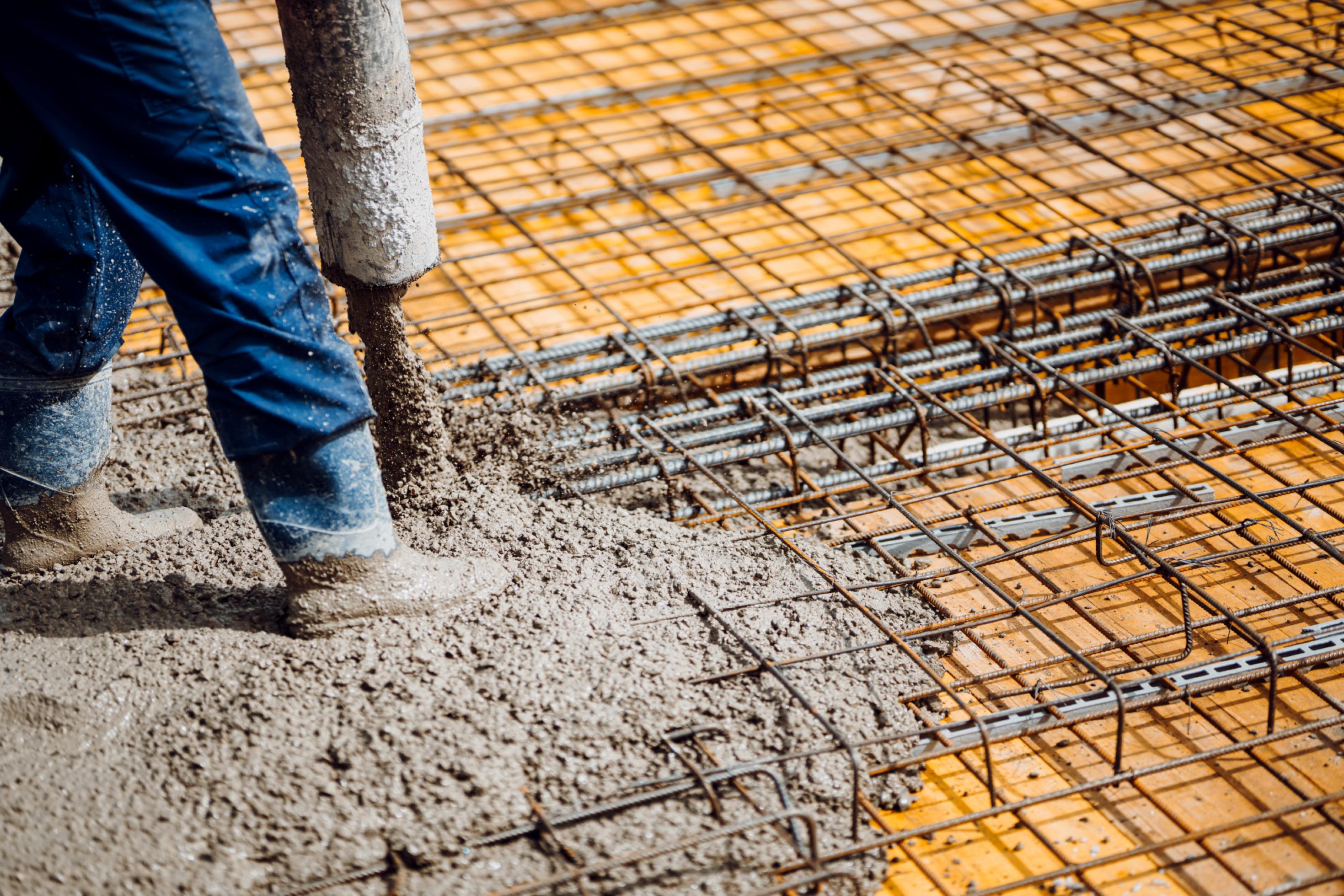Die Beine eines Menschen in Gummistiefeln stehen auf einem Geflecht aus Baustahl, während aus einem Rohr flüssiger grauer Beton daneben gegossen wird. Es ist dreckig.