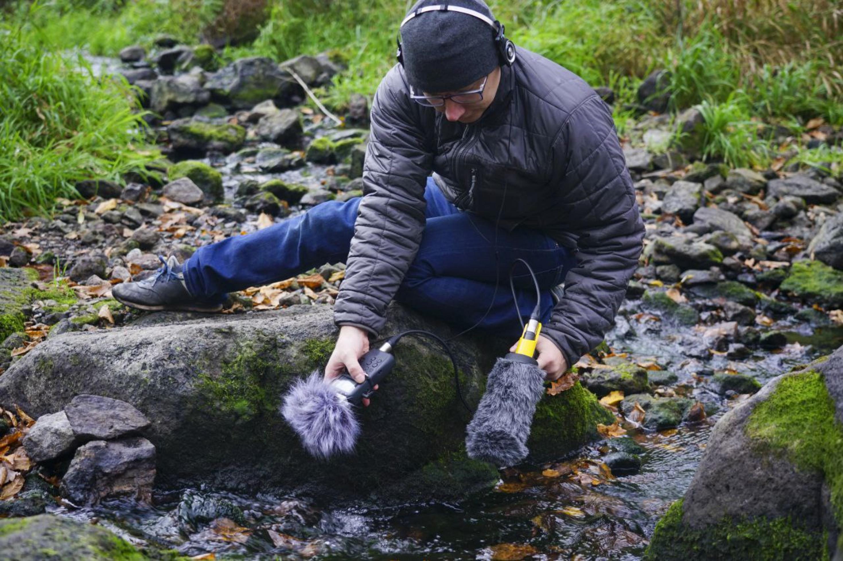Ein Mann kniet an einem Bach und hält ein Aufnahmegerät in der Hand