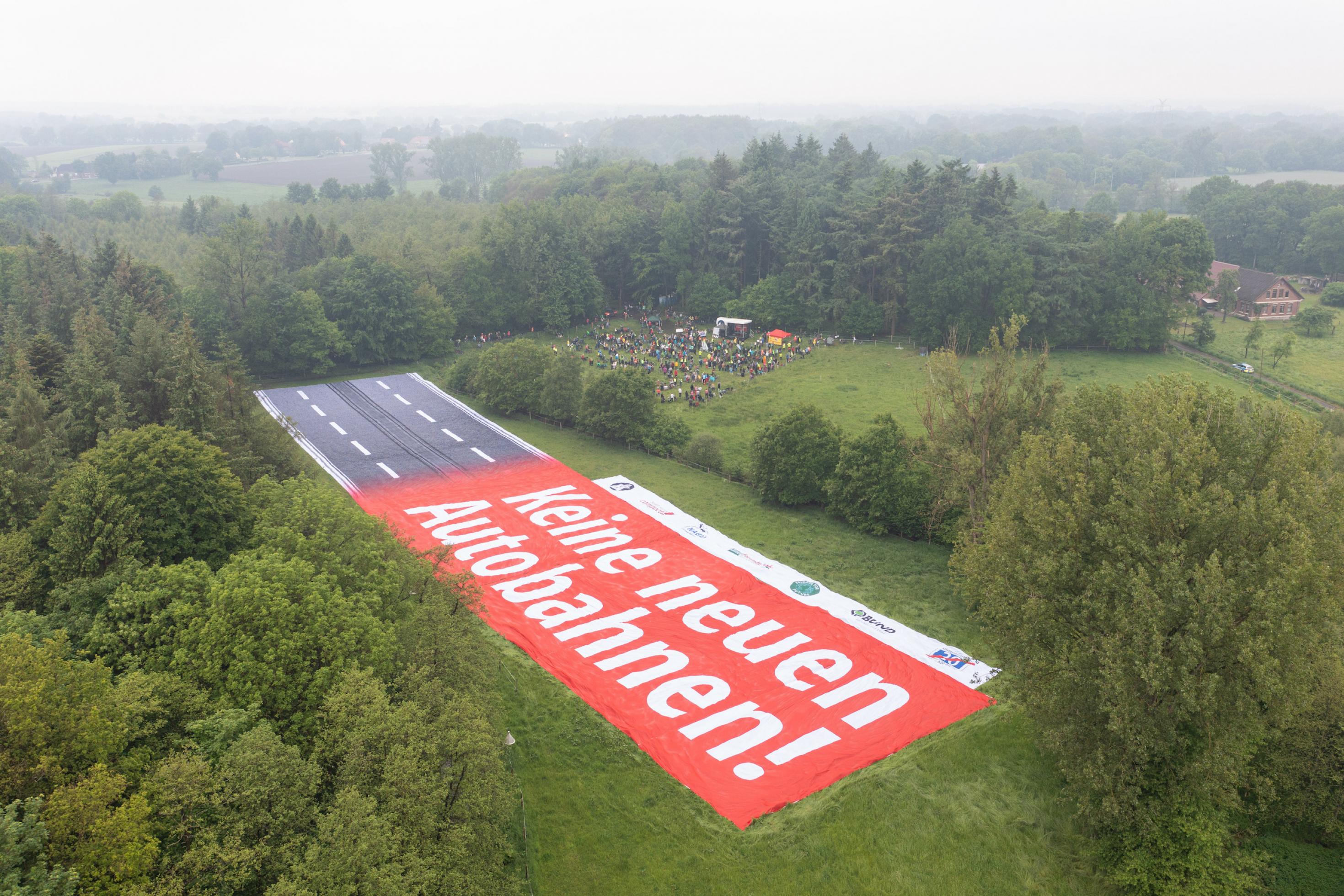 Ein Banner mit der Aufschrift „Keine neuen Autobahnen!“, der einen Autobahnabschnitt darstellt, ist auf einer Landfläche zu sehen. Es findet ein Protest gegen den geplanten Bau der A20 statt (Aufnahme mit einer Drohne).