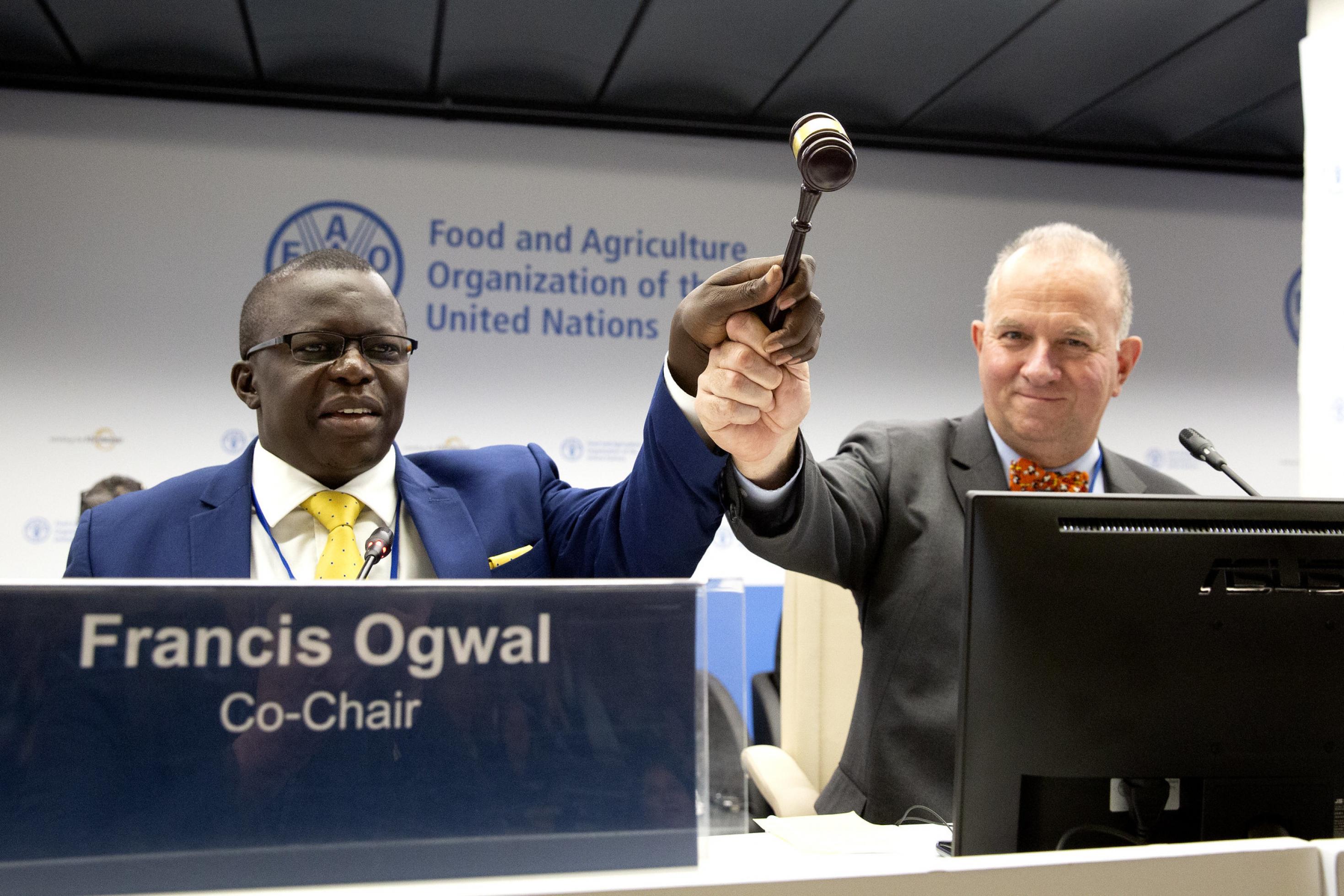 Francis Ogwal und Basil van Havre auf einem Konferenzpodium. Beide legen ihre Hände um einen Holzhammer, mit dem sie eine Konferenz eröffnen.