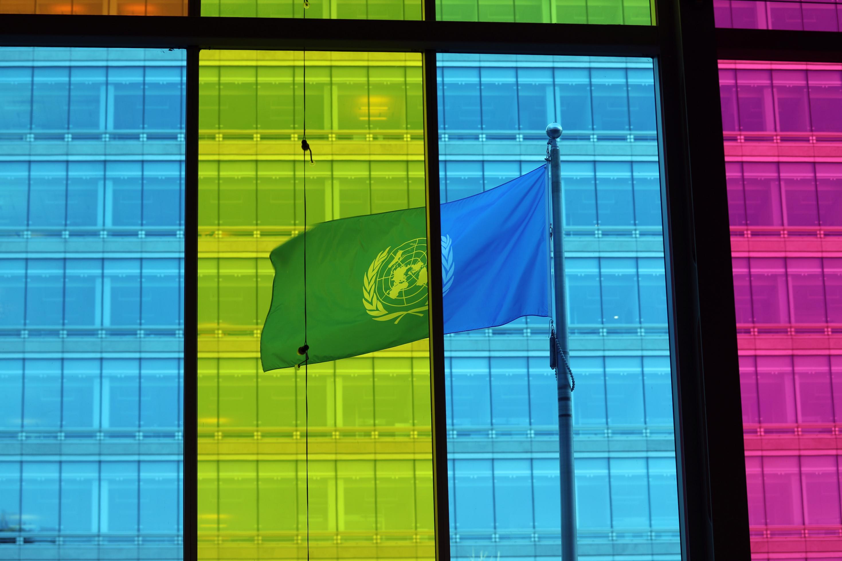 Die Fenster des Konferenzgebäudes sind in blau, gelb und lila gefärbt. Dahinter weht die UN-Fahne.