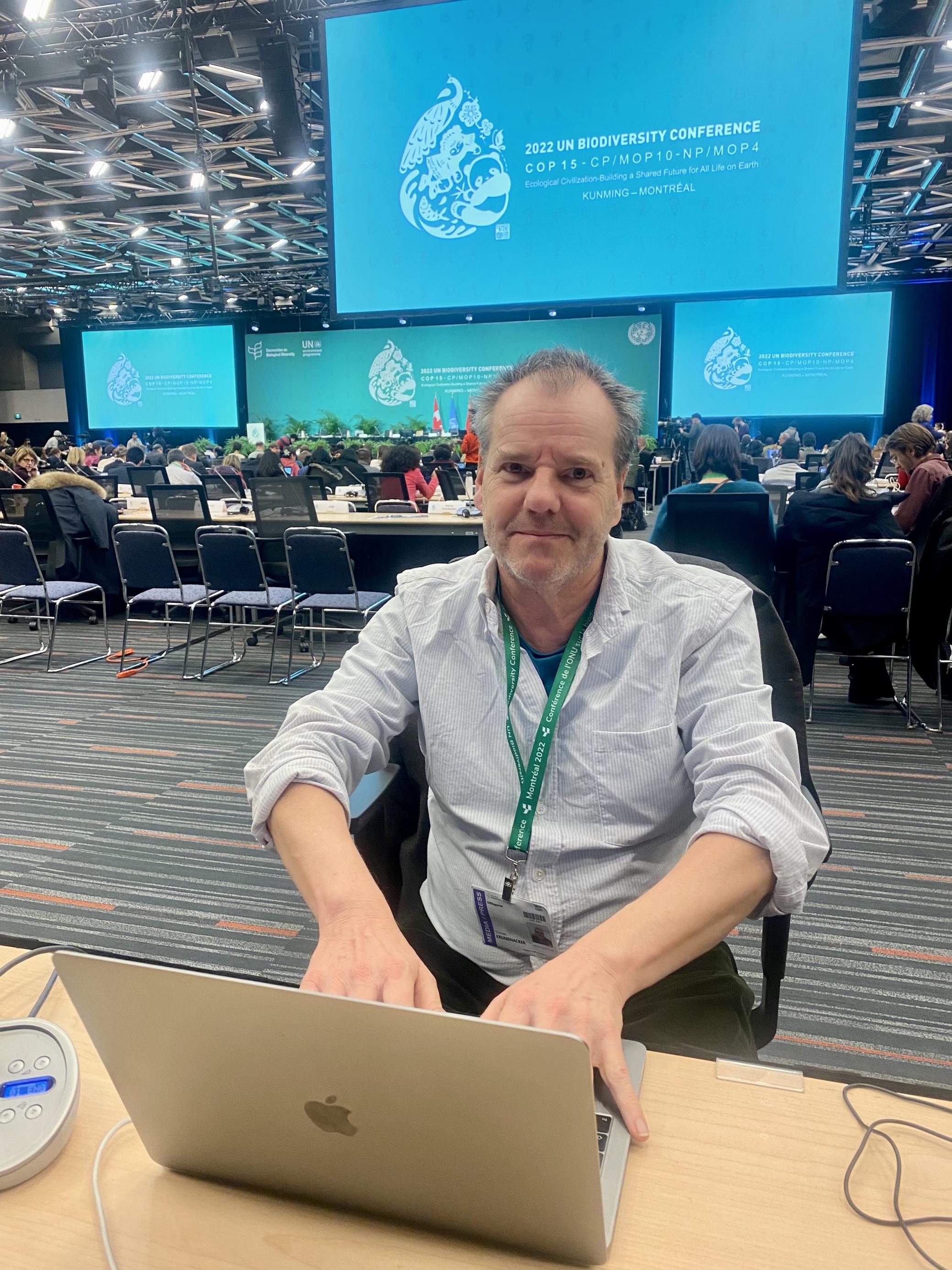 Thomas sitzt am Laptop in einer großen Veranstaltungshalle mit hellblauem Hintergrund.