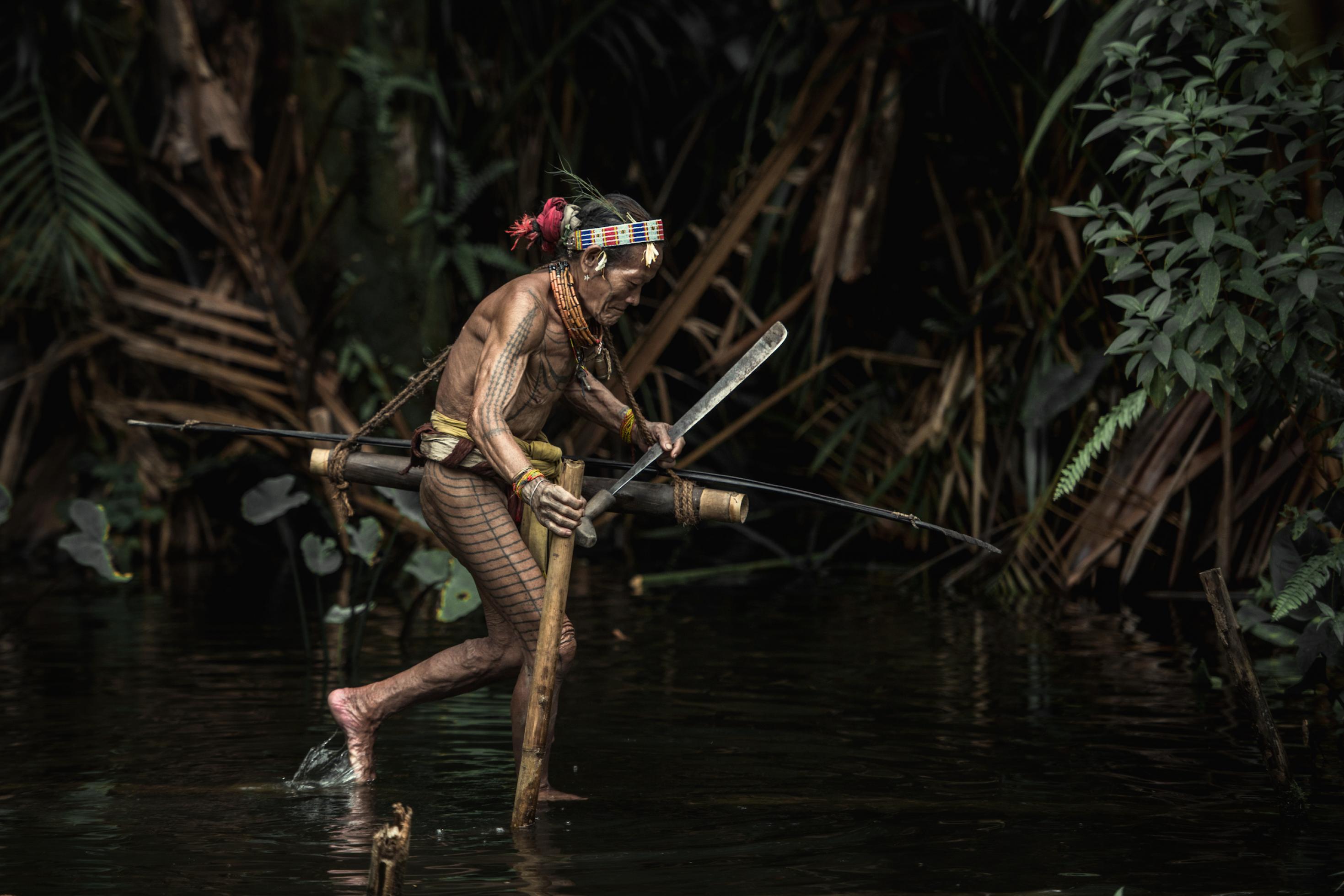 Indigener mit Tätowierungen watet durch einen Bach, er hat eine Machete in der Hand.