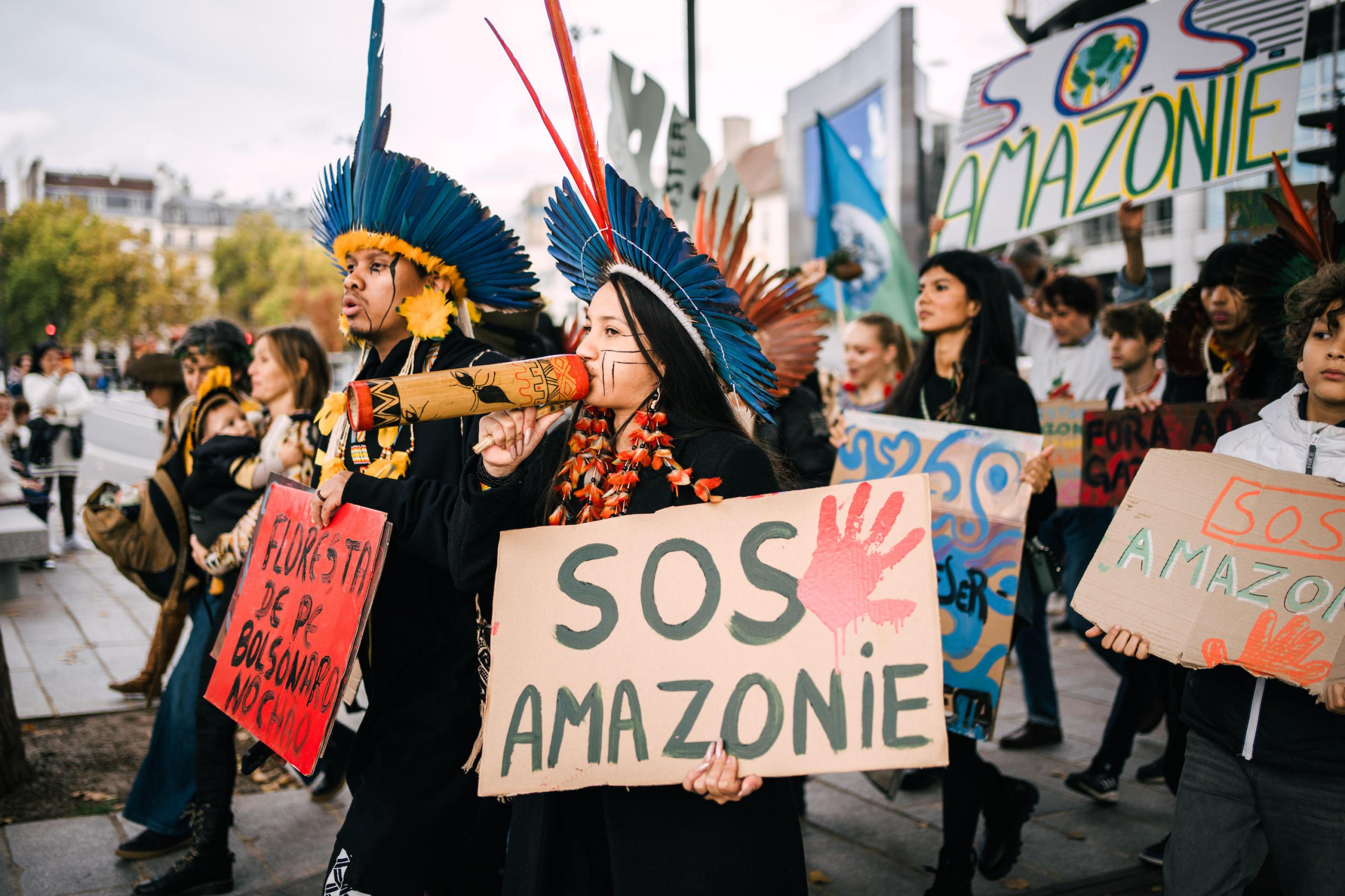 Ein Mann und eine Frau in indigenem Federschmuck führen eine Demonstration an. Auf ihren Schildern steht zum Beispiel SOS Amazonas.