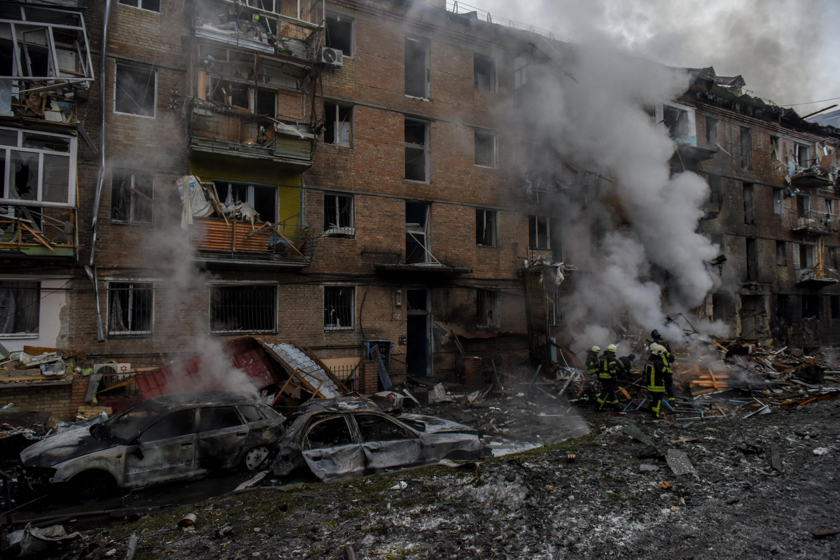 Ausgebombtes Wohngebäude, zerstörte Autos, davor Feuerwehrleute, düstere Atmosphäre.