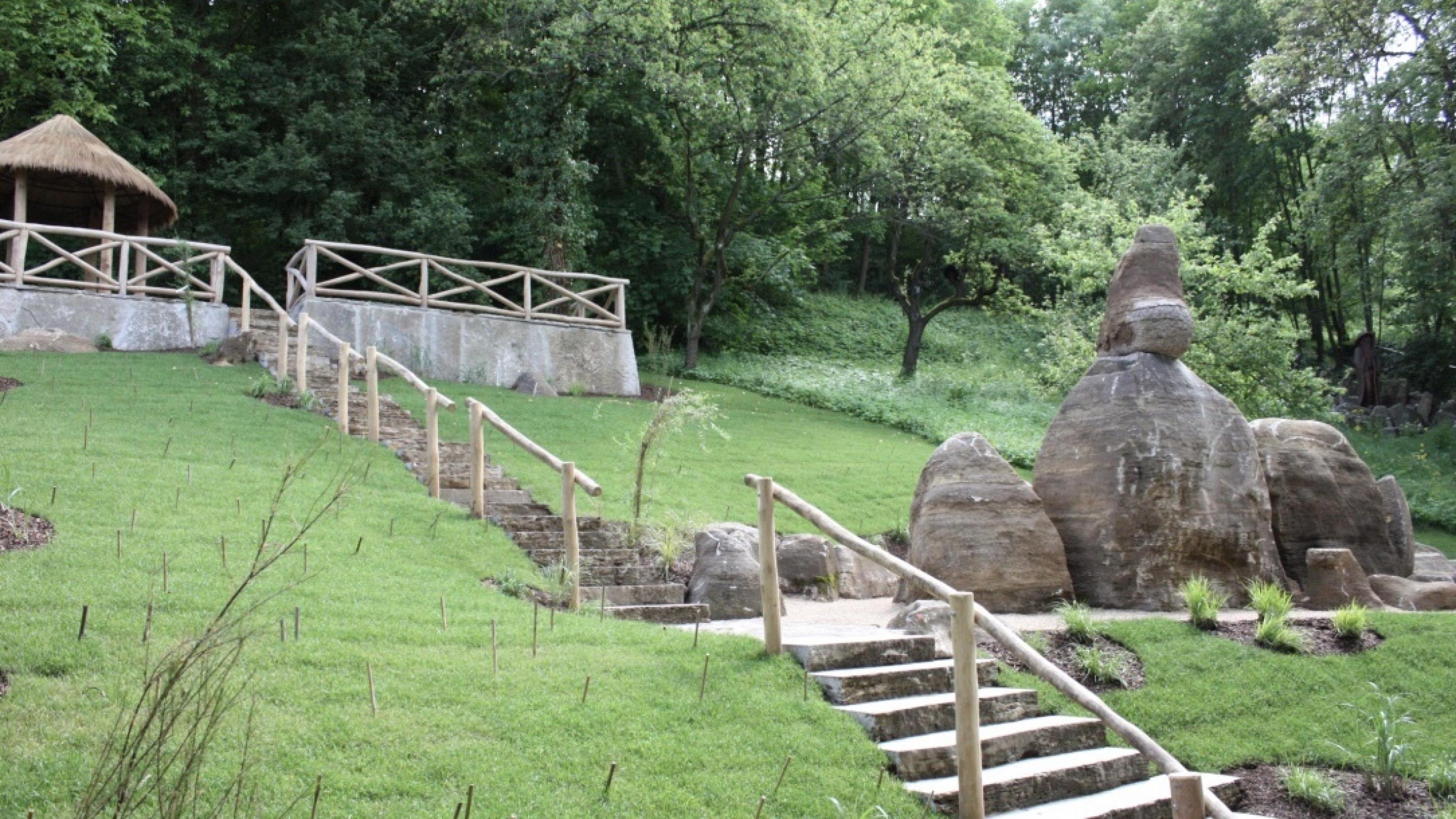 Ein parkähnlicher Garten an einem Hügel, mit grünem Rasen. Eine Treppe führt hinauf zu einer „afrikanischen“ Hütte, welche links oben auf dem Bild zu sehen ist. Rechts im Bild eine Felsformation, welche die Formen des Bismarck Rock in Mwanza hat.