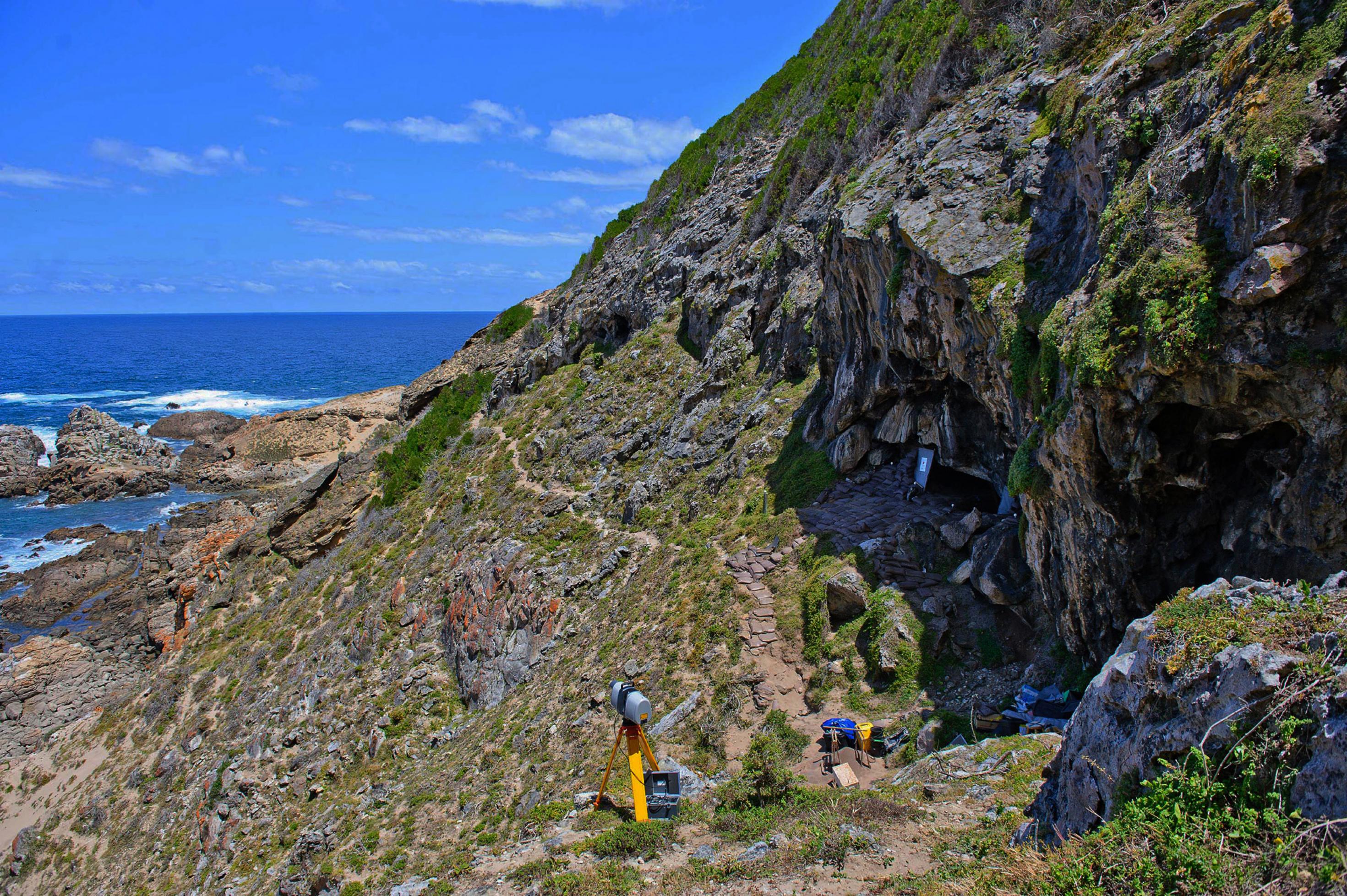 Auf der rechten Bildseite sieht man einen steil ansteigenden Berg, vorne rechts den Eingang zu einer Höhle, der oben von Felsen überwölbt wird. Vor der Höhle stehen wissenschaftliche Messgeräte. Ein Trampelpfad führt von der Höhle fort, den Hang entlang. Links fällt das Gelände steil ab bis zum Meer, das unten an Felskuppen brandet. Darüber spannt sich ein blauer Himmel mit wenigen, kleinen Wolken.