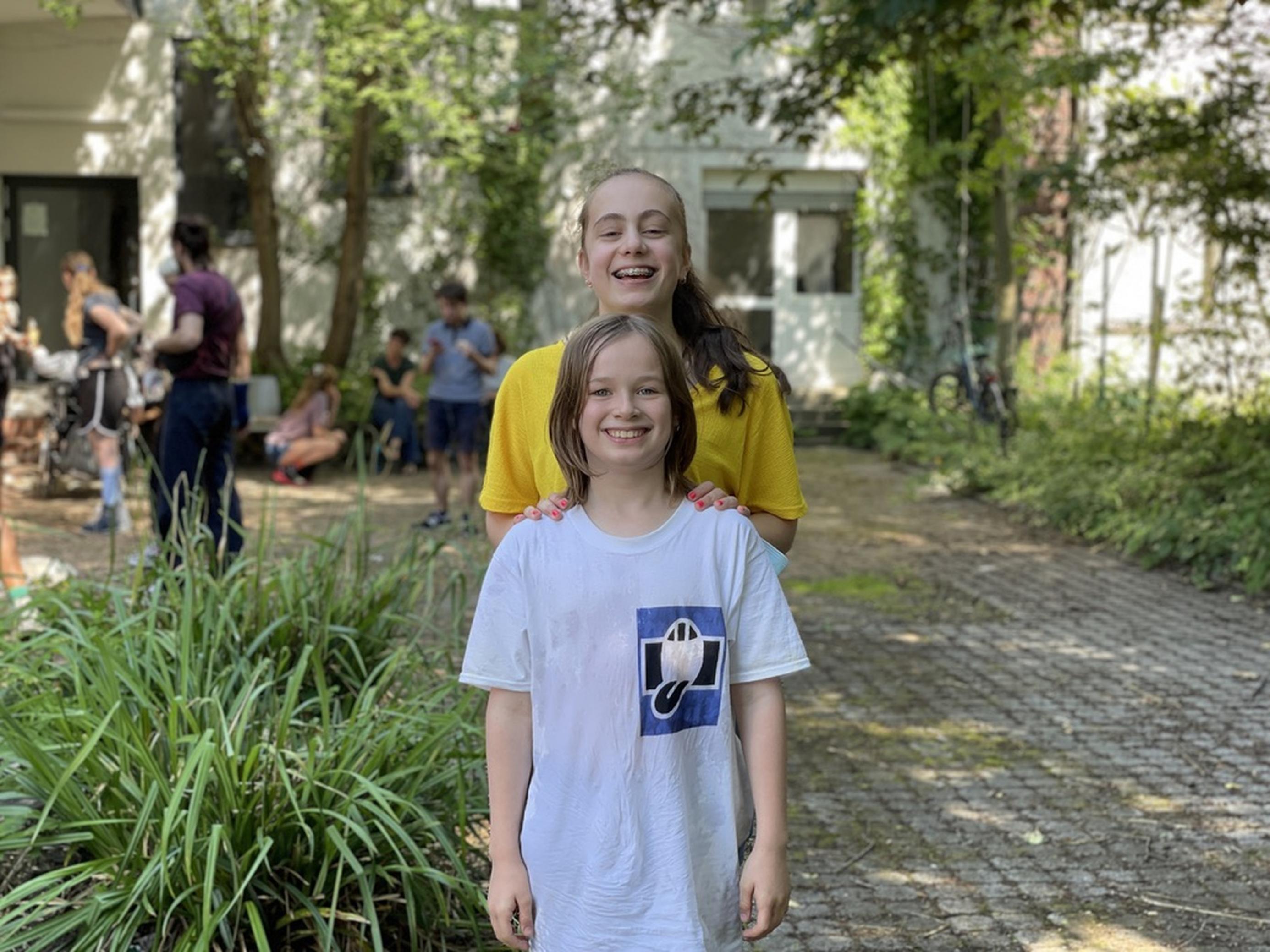 Ariane (14) steht im begrünten Hof hinter Henri (11) und legt ihm die Hände auf die Schulter. Beide lachen in die Kamera.