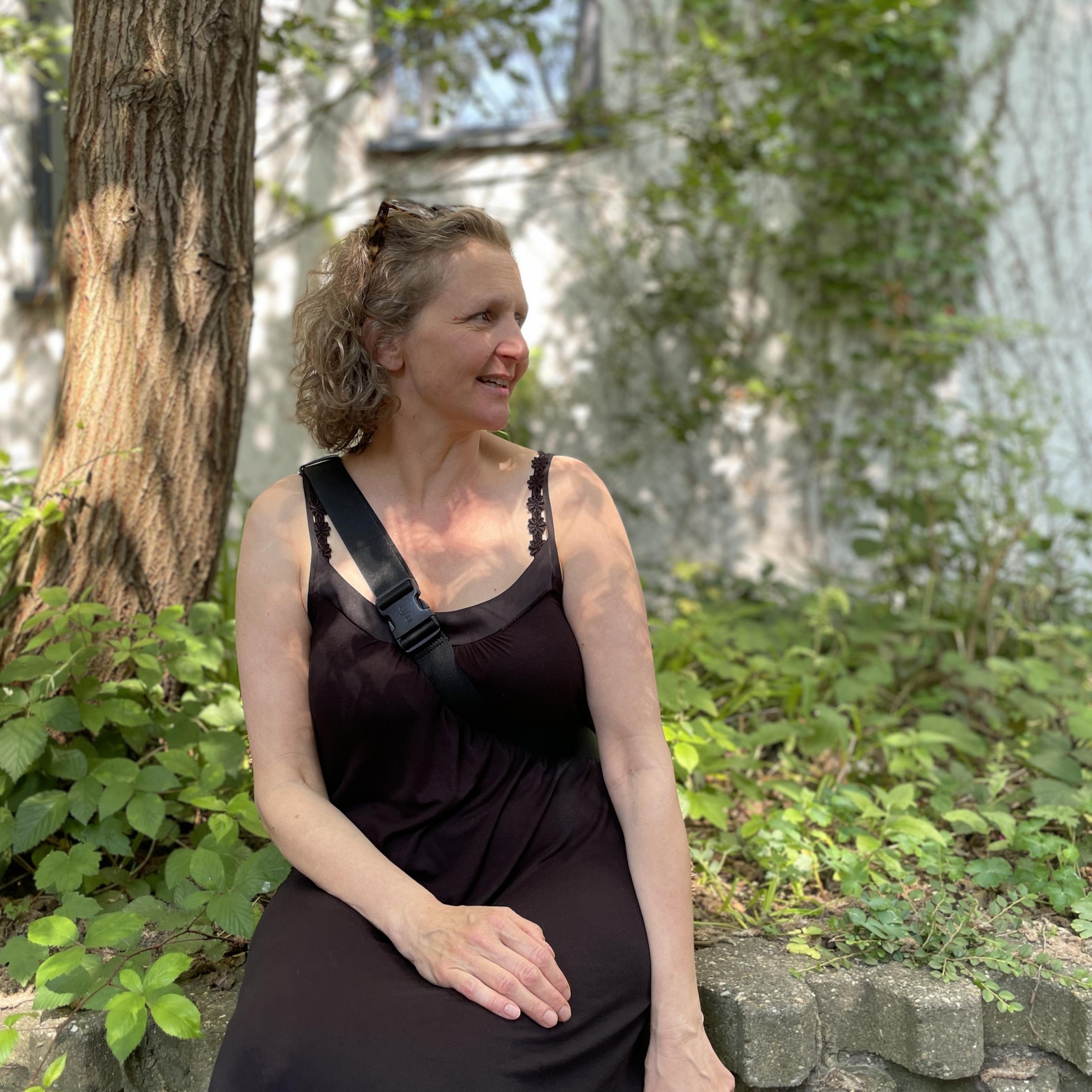 Cathrin Rose sitzt im schwarzen Kleid auf einem Mäuerchen unter einem Baum und blinzelt in die Sonne.