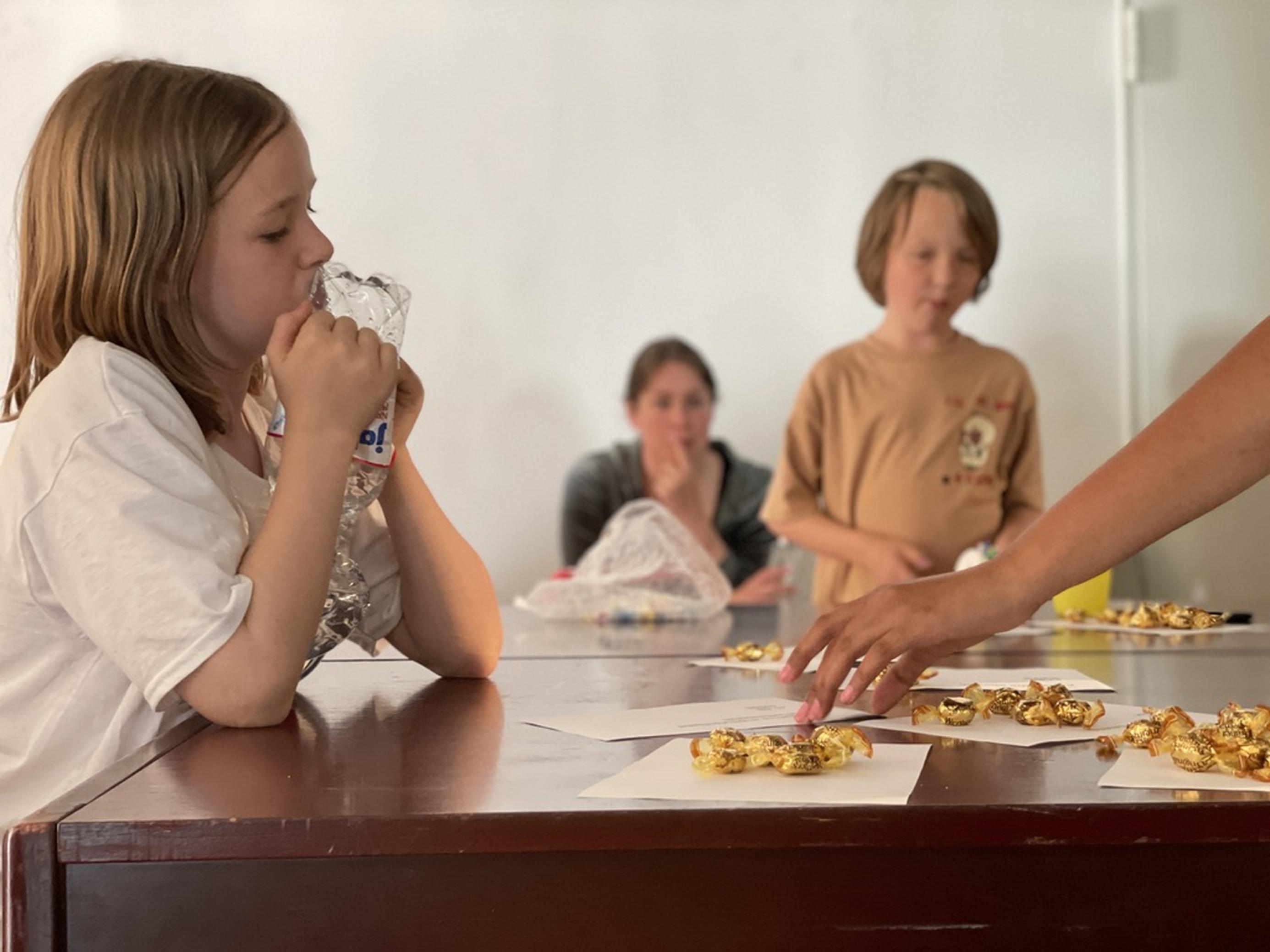 Henri sieht Alicia beim Verteilen der Bonbons, also Geldbeträge, zu. Im Hintergrund sieht man Henri (8) am Tisch und dahinter eine Teilnehmerin aus dem Workshop, die den Jugendlichen zusieht.