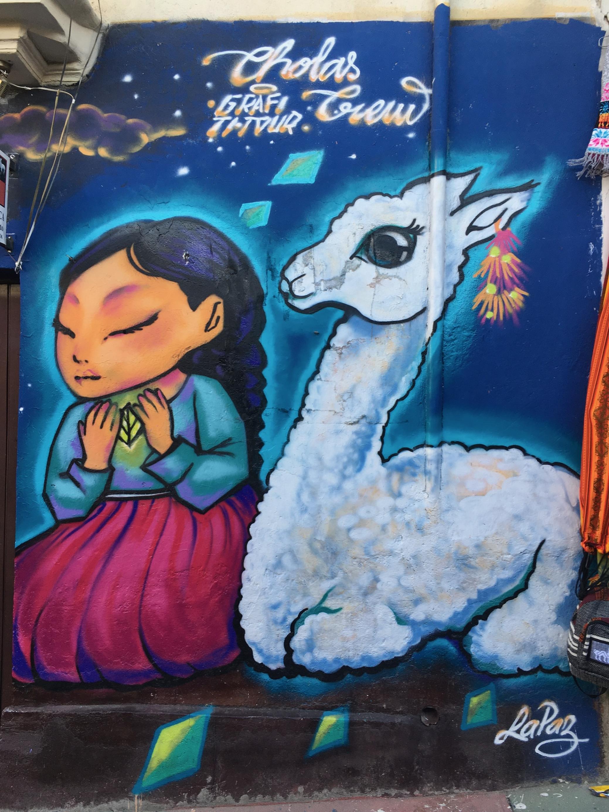 Bunte Wandmalerei zeigt indigene Frau mit geschlossenen Augen und langen Zöpfen, die ein Blatt in den Händen trägt. Neben ihr sitzt ein schneeweißes Lama.