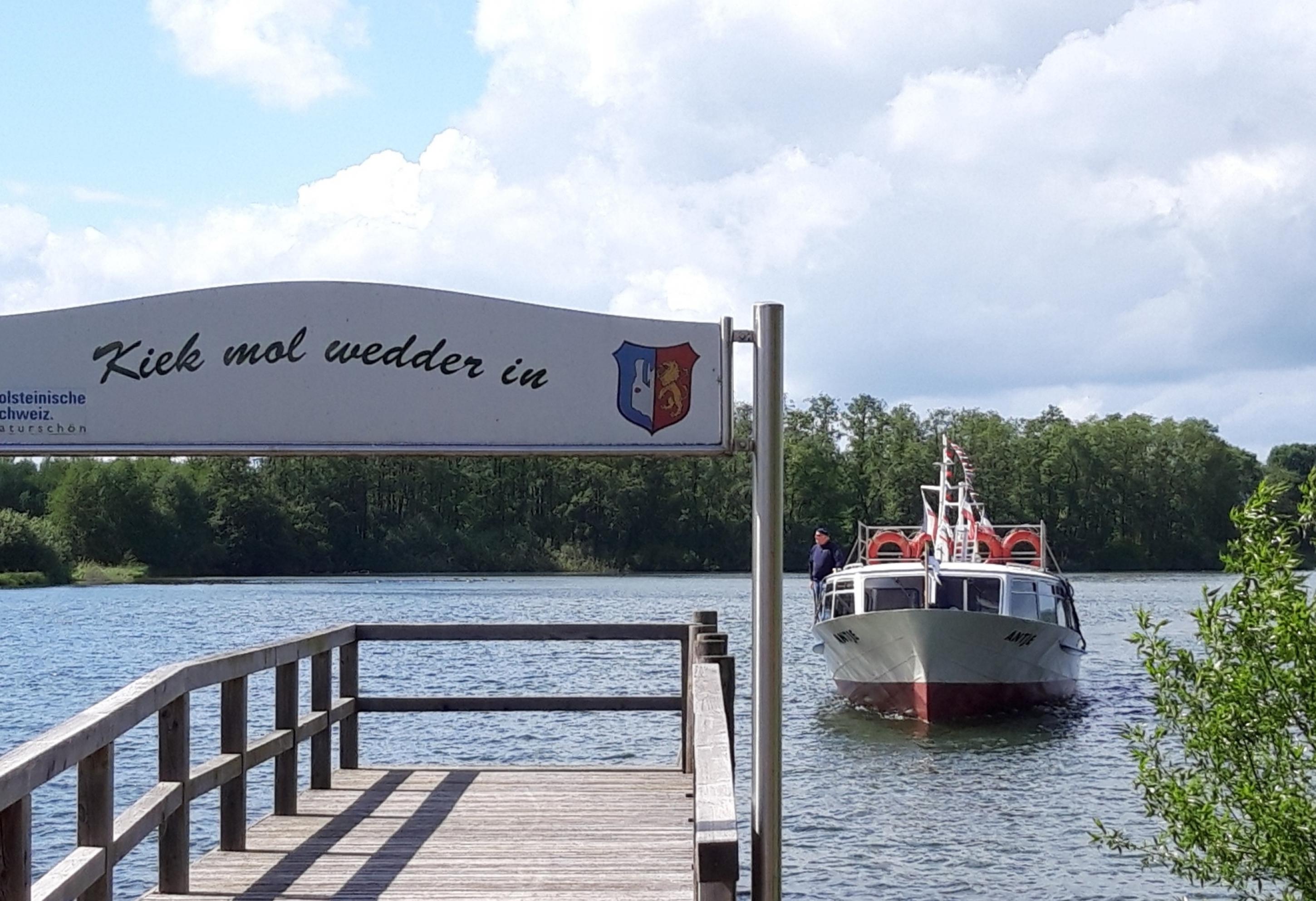 „Kiek mal wedder in“ heißt es auf dem den Bootsanleger überspannenden Holzpanel mit dem Wappen des Ortes Bosau, den soeben ein Schiff ansteuert.