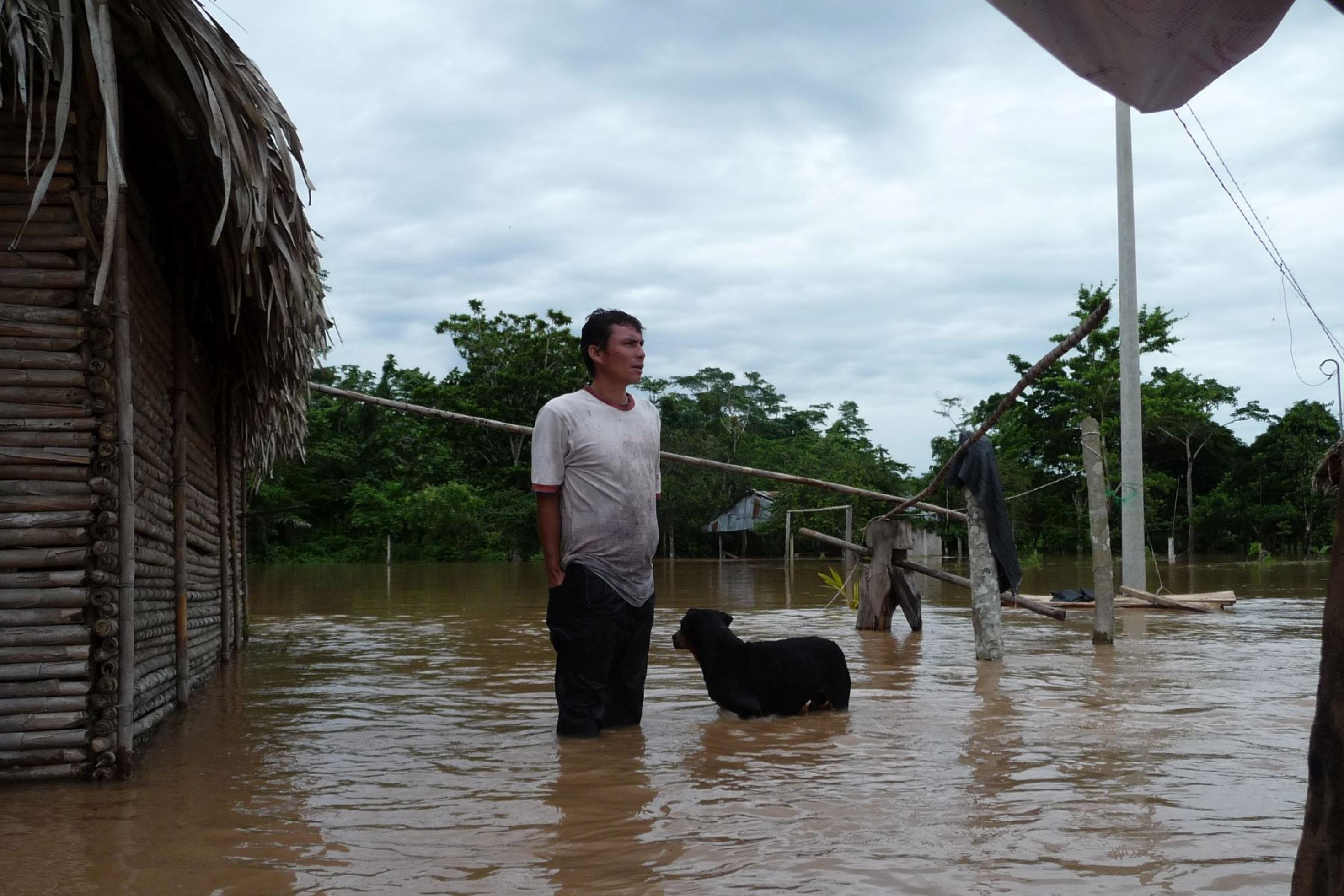 Mann und Hund stehen bis zu den Knien im Wasser, daneben ein einfaches Holzhaus mit Palmdach