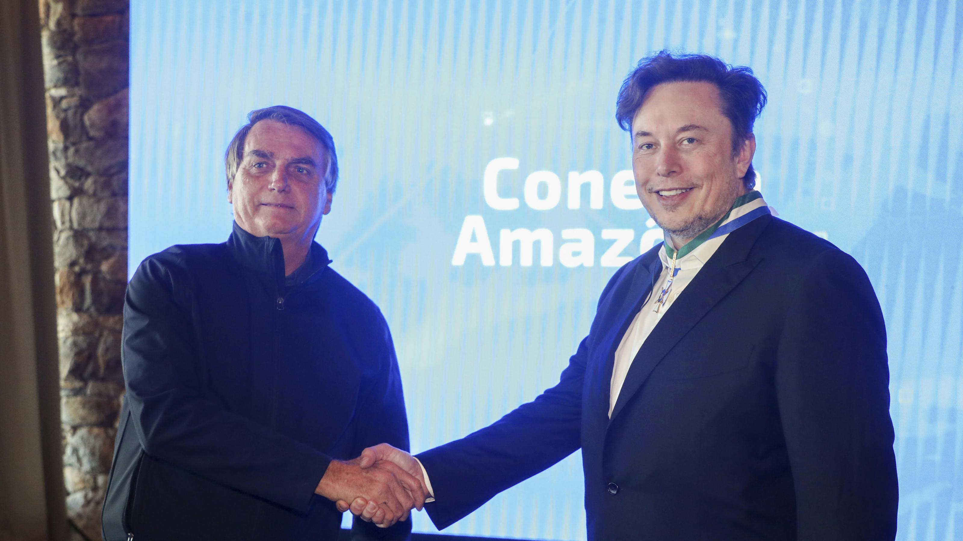 Jair Bolsonaro und Elon Musk geben sich die Hand vor einem blauen Hintergrund in einem Konferenezzentrum.