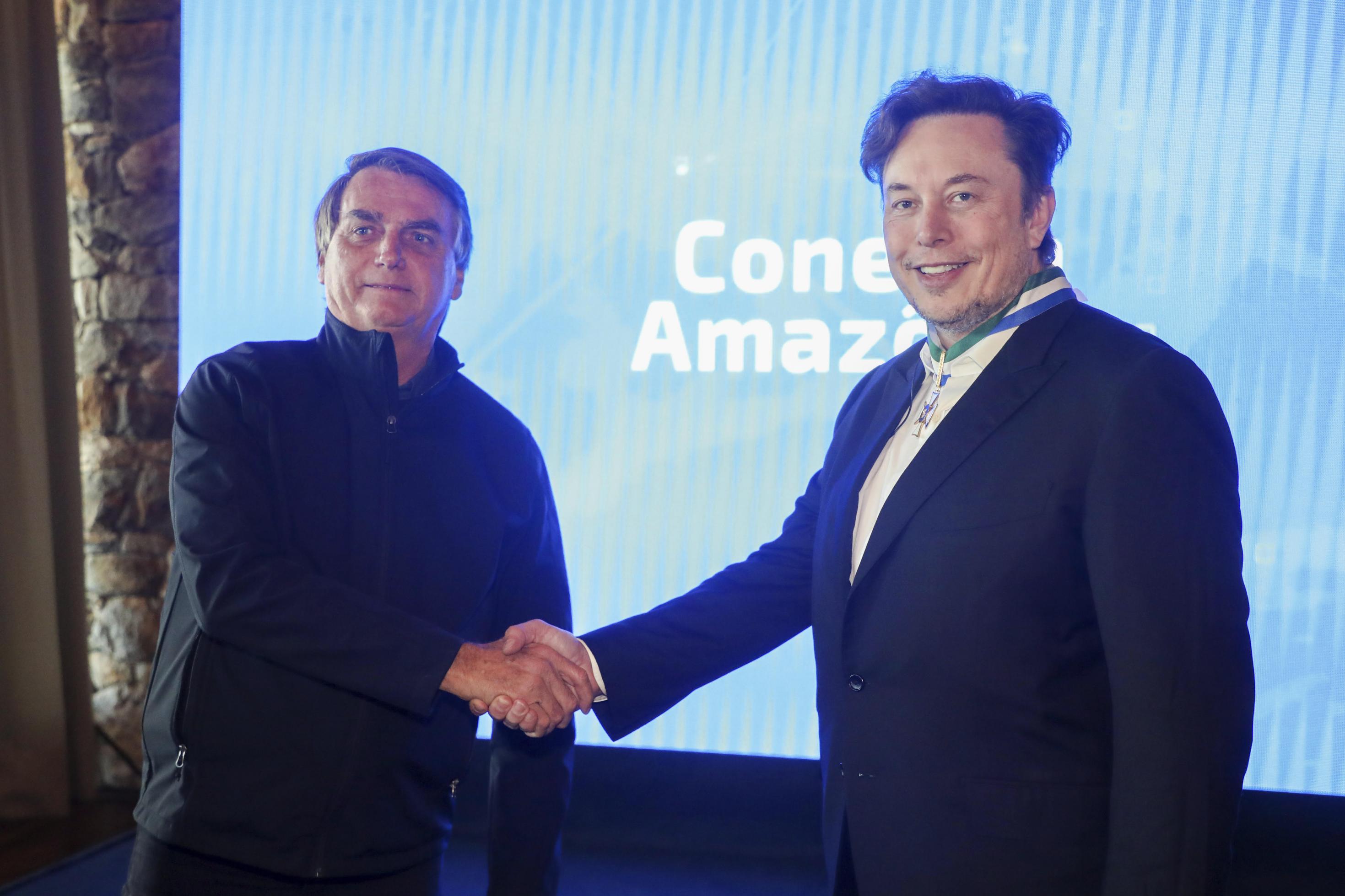 Jair Bolsonaro und Elon Musk geben sich die Hand vor einem blauen Hintergrund in einem Konferenezzentrum.