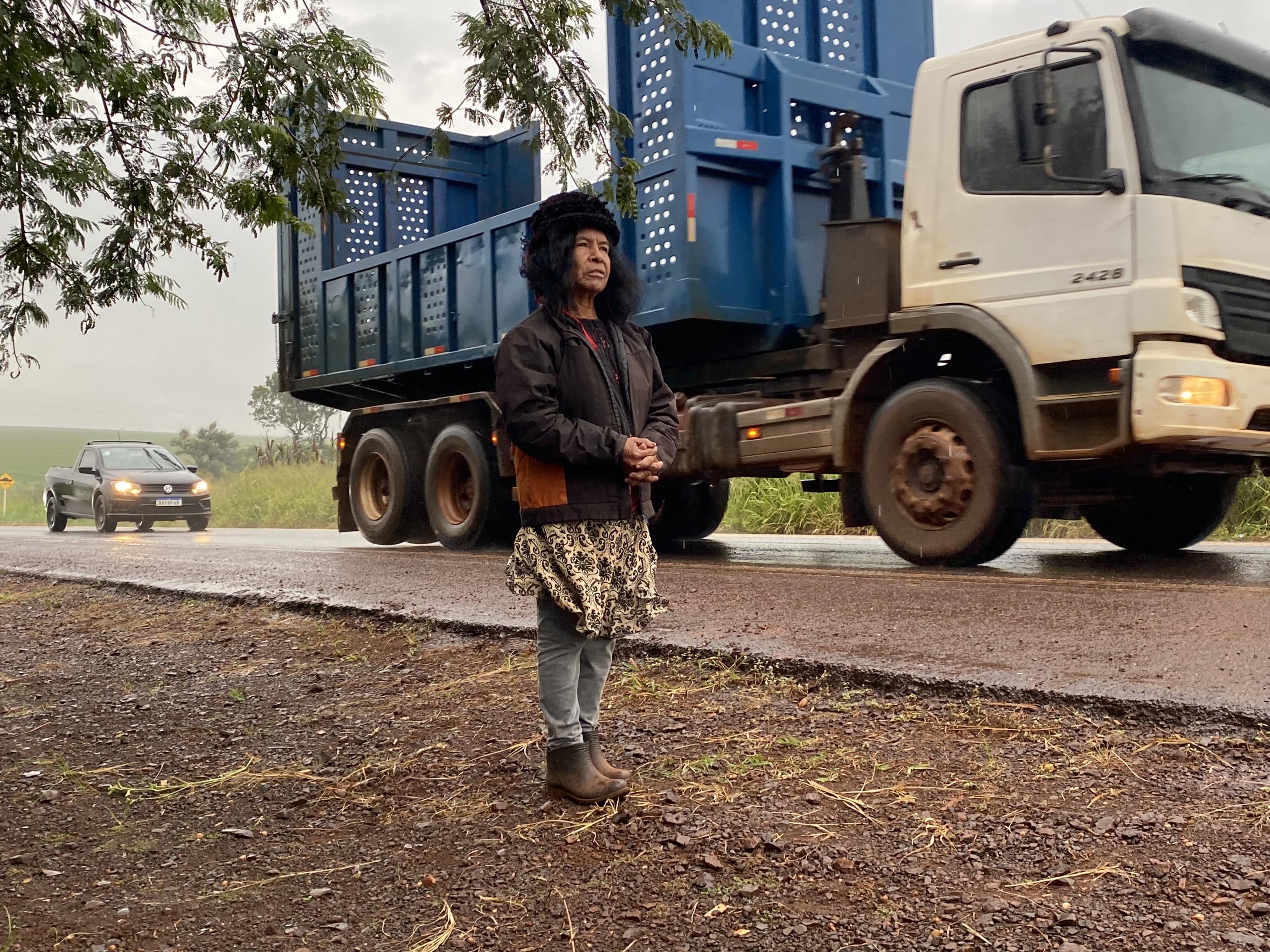 Eine indigene Frau wartet am StrEine indigene Frau wartet am Strassenrand des Sojahighways in Südbrasilien, während Schwerlaster vorbeibrausen.