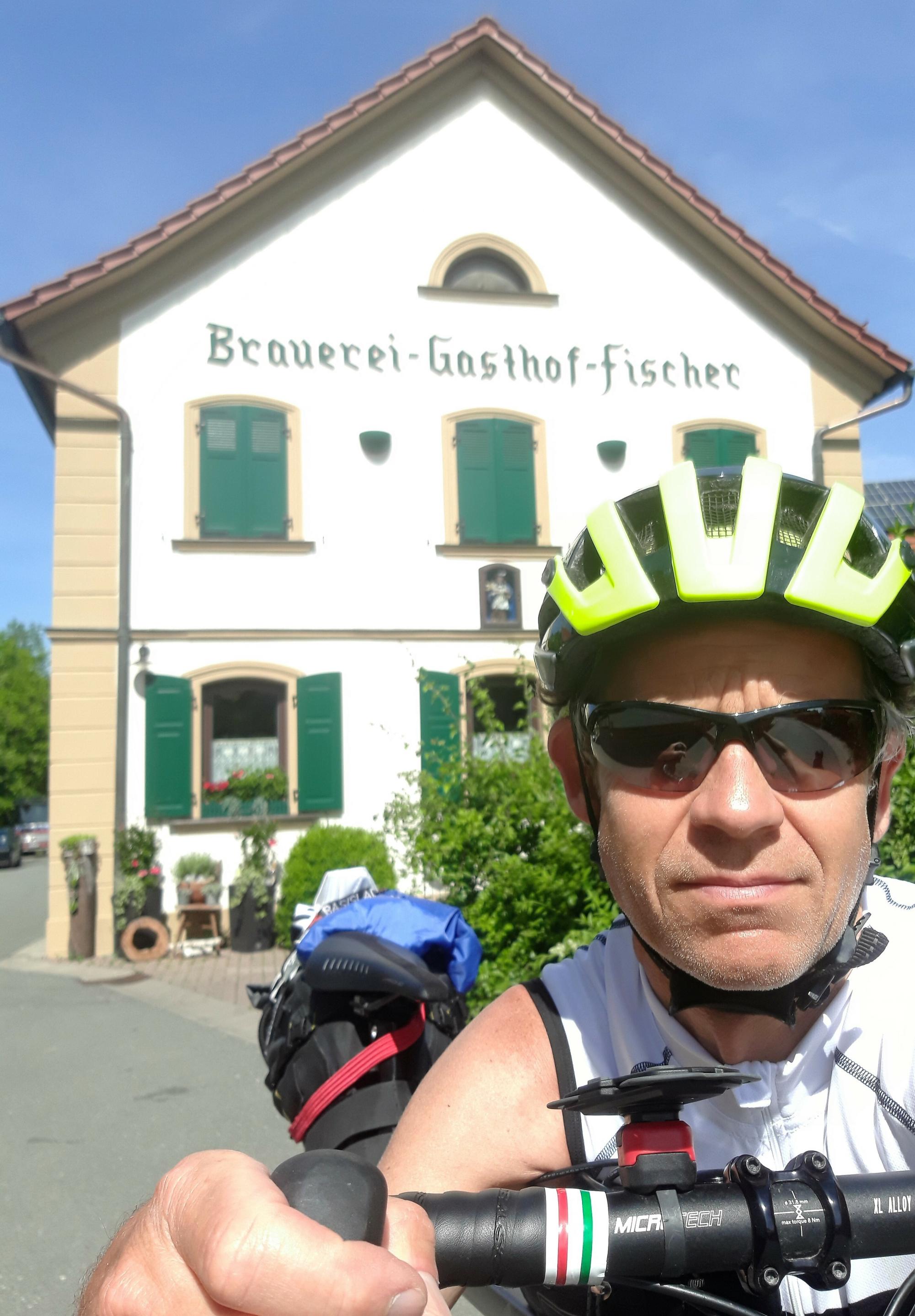 Selfie des Helm-bewehrten RadelndenReporters vor der weißen Hausfront der Brauerei, Aufschrift dort: Brauerei-Gasthof-Fischer.