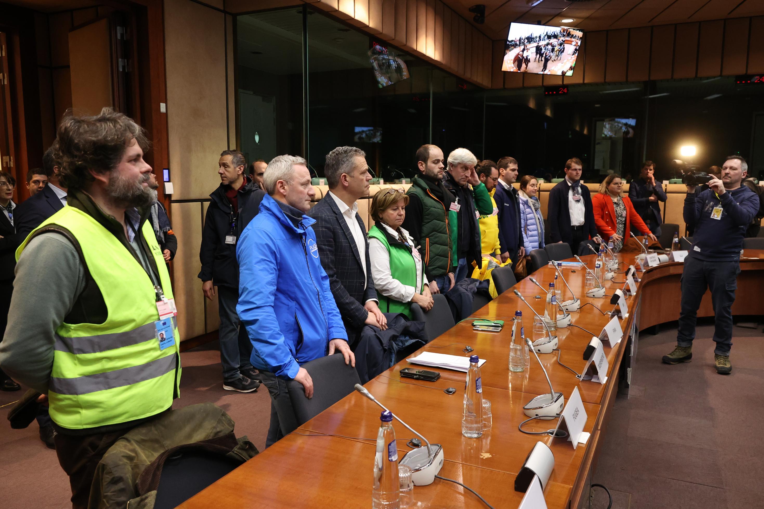 Bäuerinnen und Bauern, der erst in gelber Warnweste, stehen vor Tischen im EU Parlament. Es wirkt, als hätten sie sich zur Begrüßung von ihren Plätzen erhoben.