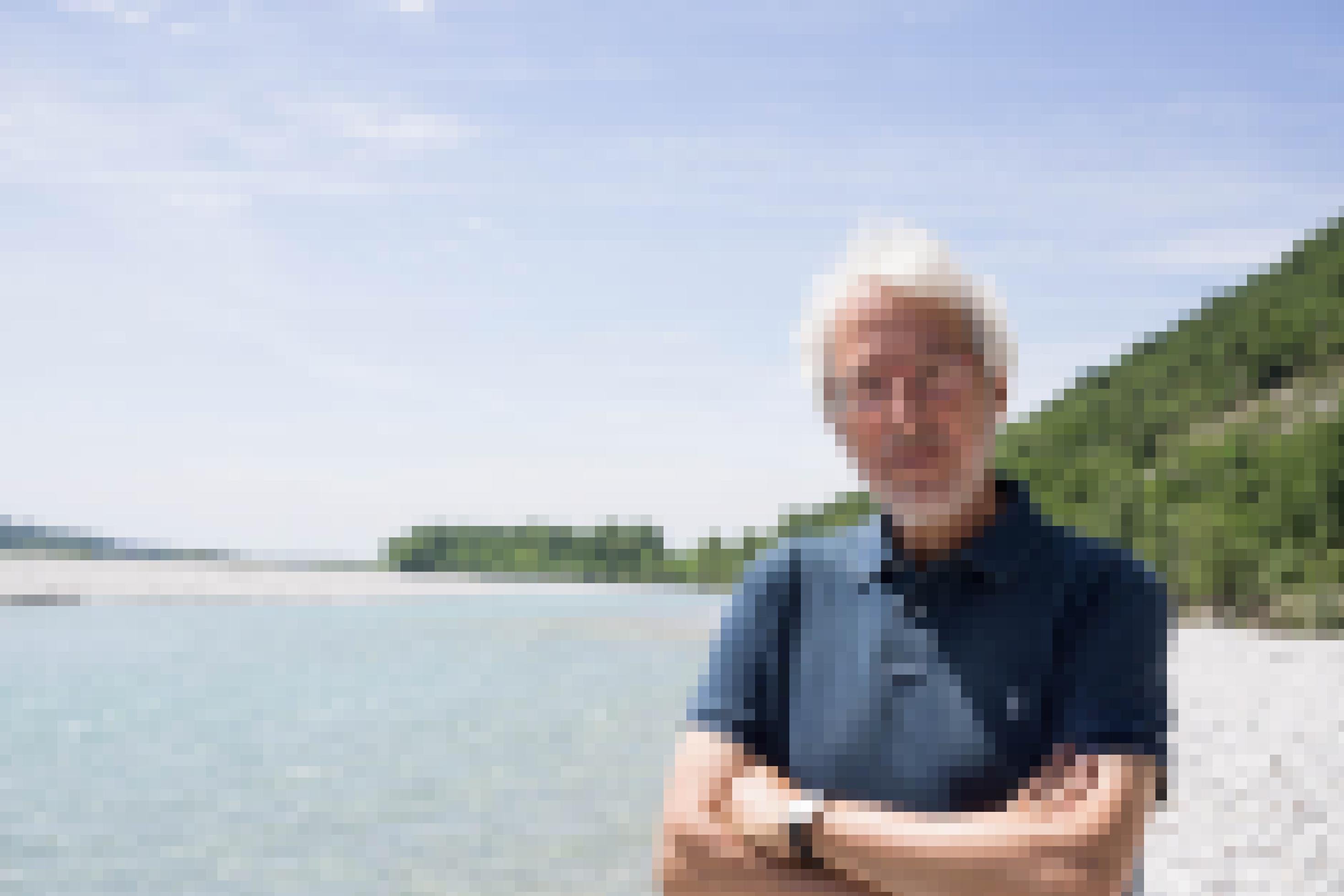 Mann mit weißem Haar und Vollbart steht an Fluss mit viel Schotter und türkisfarbenem Wasser.
