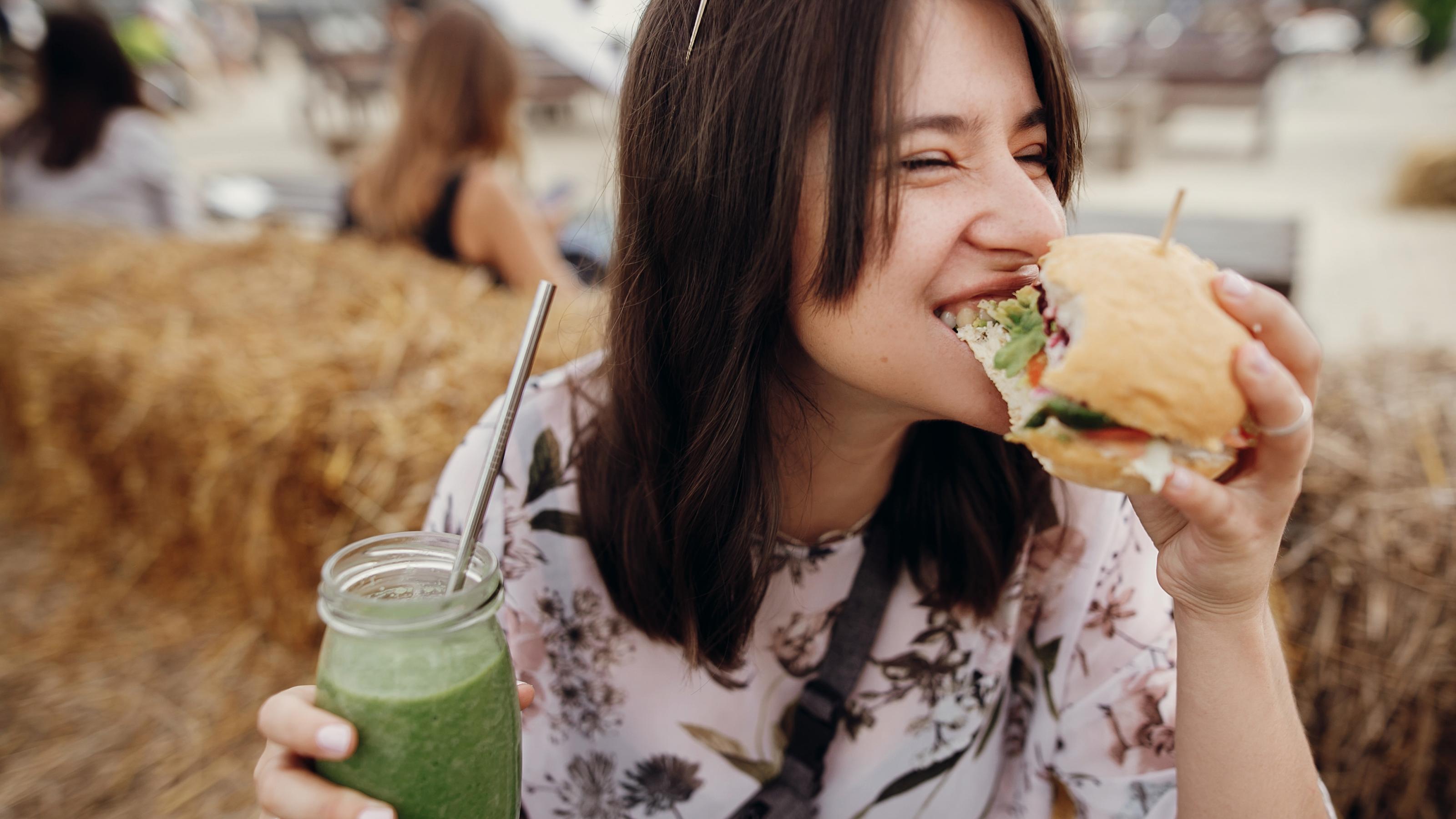 Eine junge Frau mit braunen Haaren hält in einer Hand ein Glas gefüllt mit einem grünen Smoothie, in der anderen hält sie einen vegetarischen Burger, in den sie herzhaft reinbeißt.