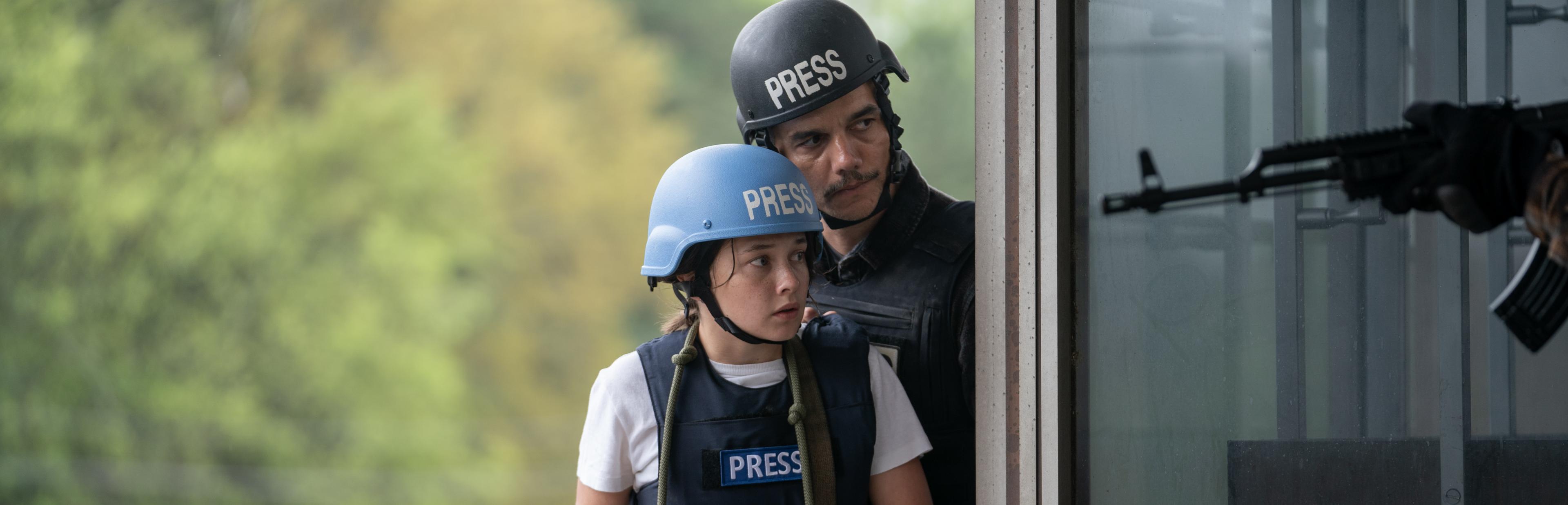 Eine junge Reporterin und ein älterer Kollege tragen Helm und kugelsichere Weste. Im Vordergrund hält jemand ein Gewehr.