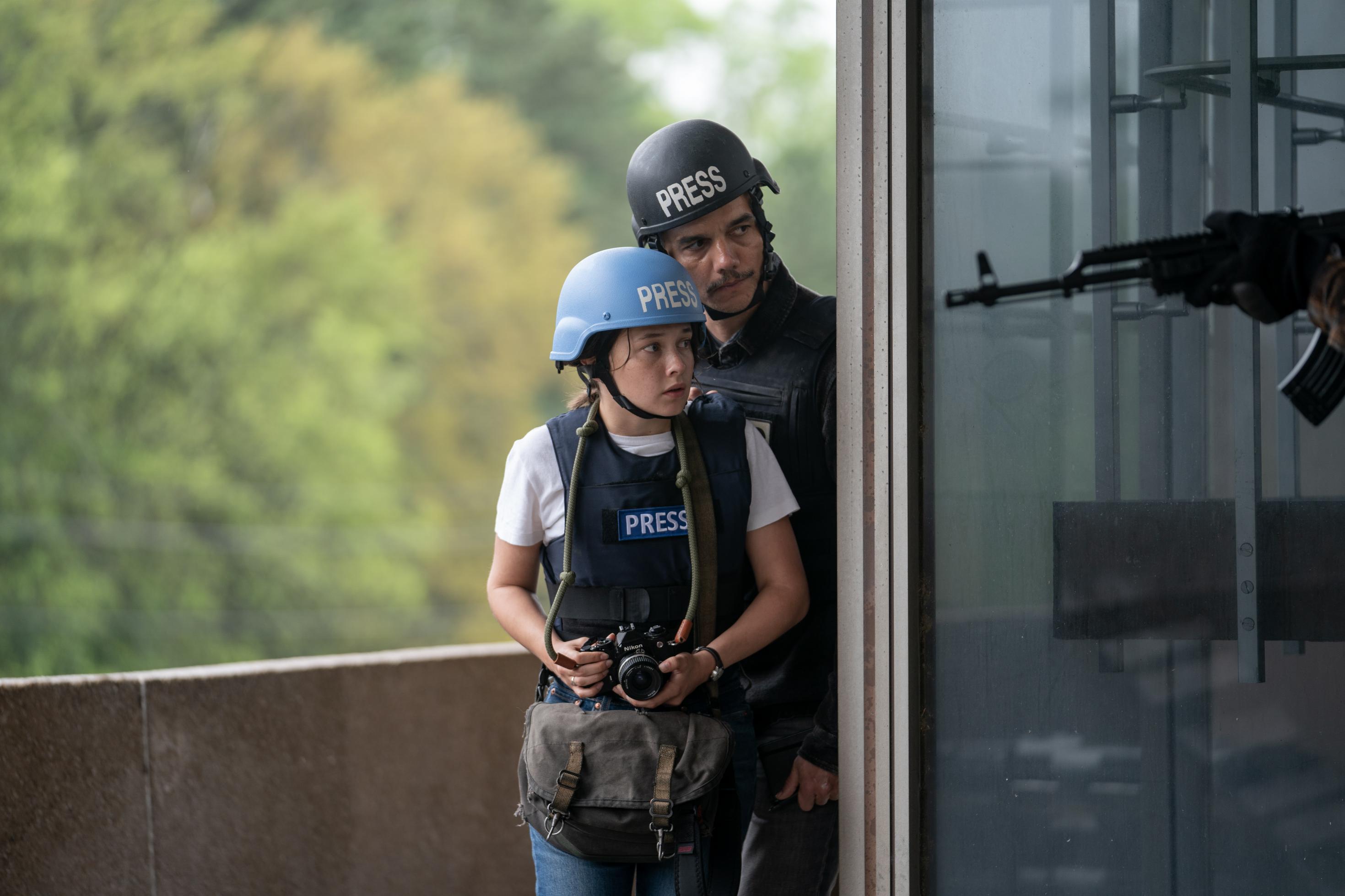 Eine junge Reporterin und ein älterer Kollege tragen Helm und kugelsichere Weste. Im Vordergrund hält jemand ein Gewehr.