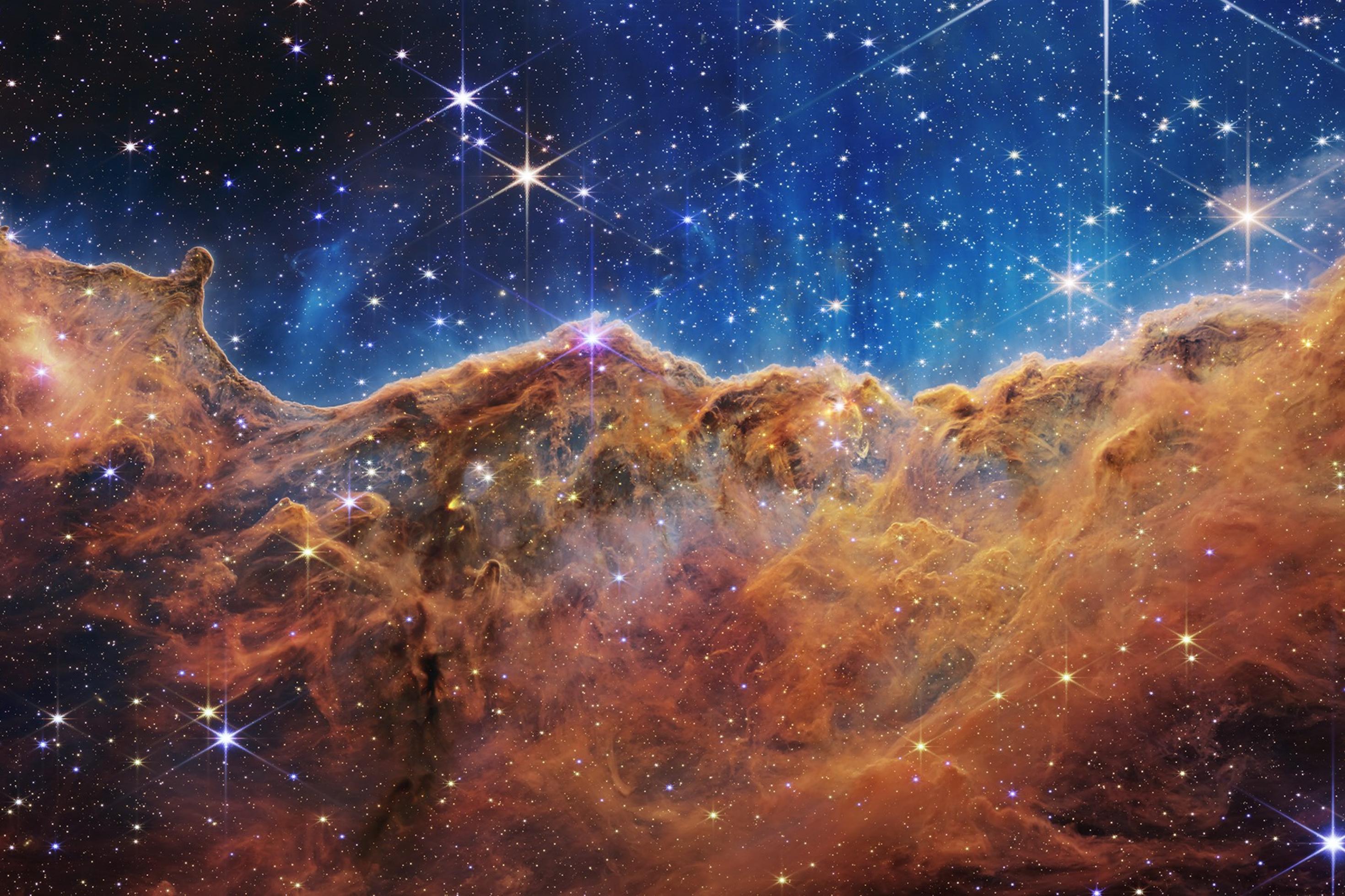 Das mit dem James Webb Space Telescope im Infraroten aufgenommene und farbcodierte Bild zeigt in der unteren Hälfte die Sternentstehungsregion im Carina-Nebel in rot; oberhalb davon leuchtet Restgas blau, im Hintergrund sind Sterne zu sehen.