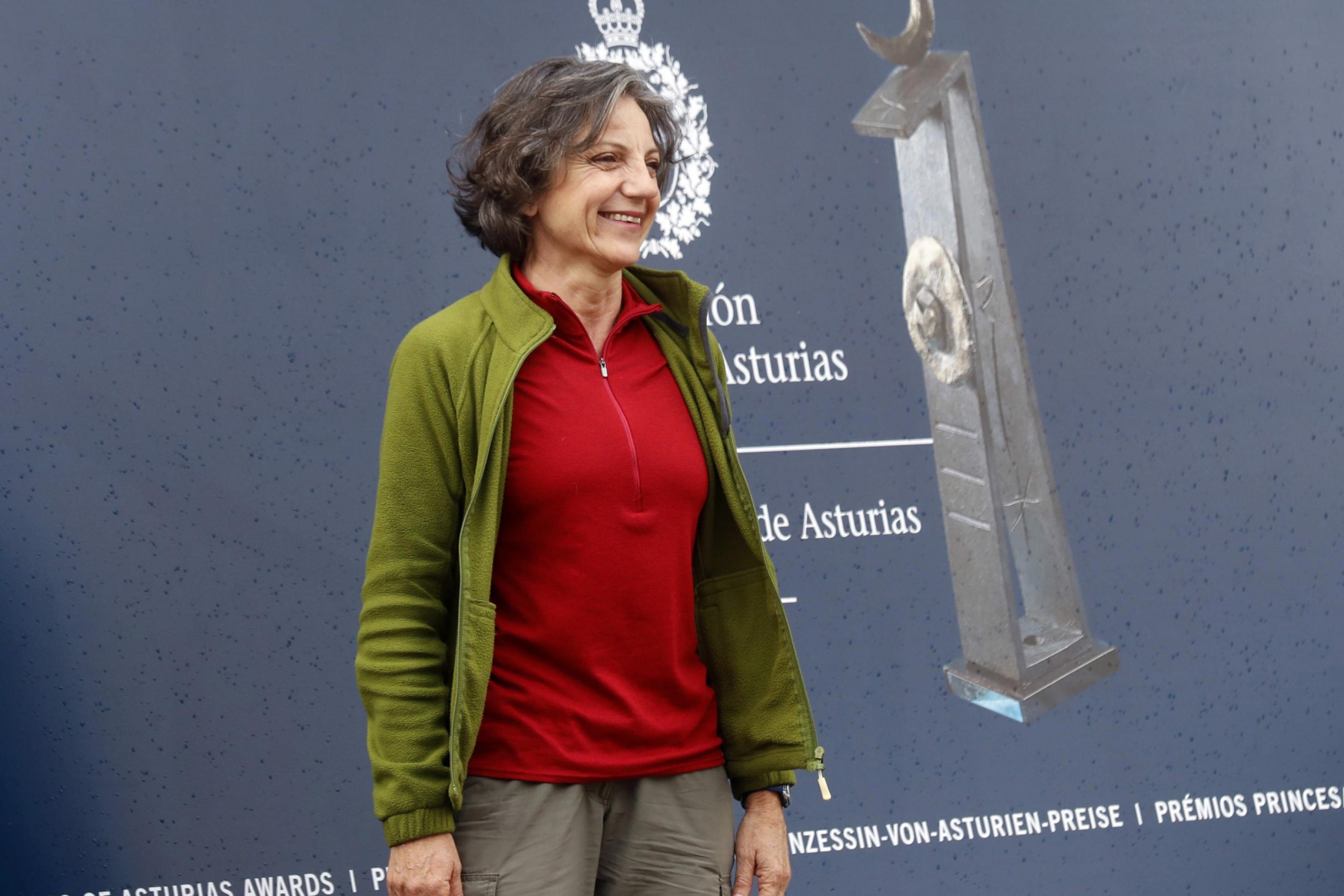 Eine mittelalte Frau mit lockigen grauen Haaren und legerer Kleidung steht vor einer grauen Wand und lacht