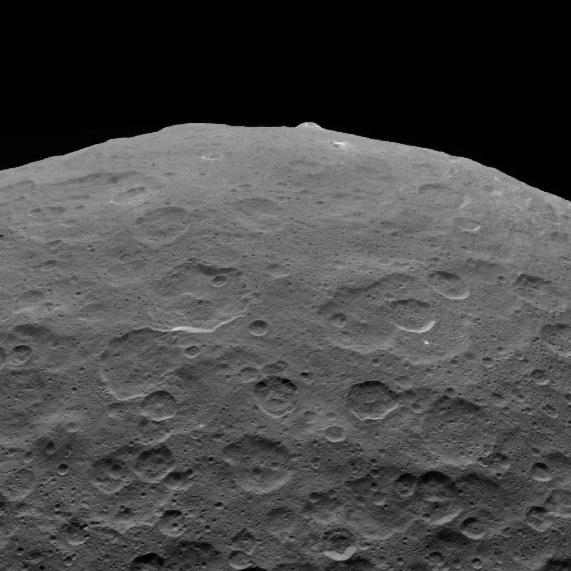 Seitliche Ansicht auf einen Teil von Ceres, wo die planetare Rundung sichtbar wird – darauf kront wie ein Pickel auf einem Gesicht der Berg Ahuna Mons