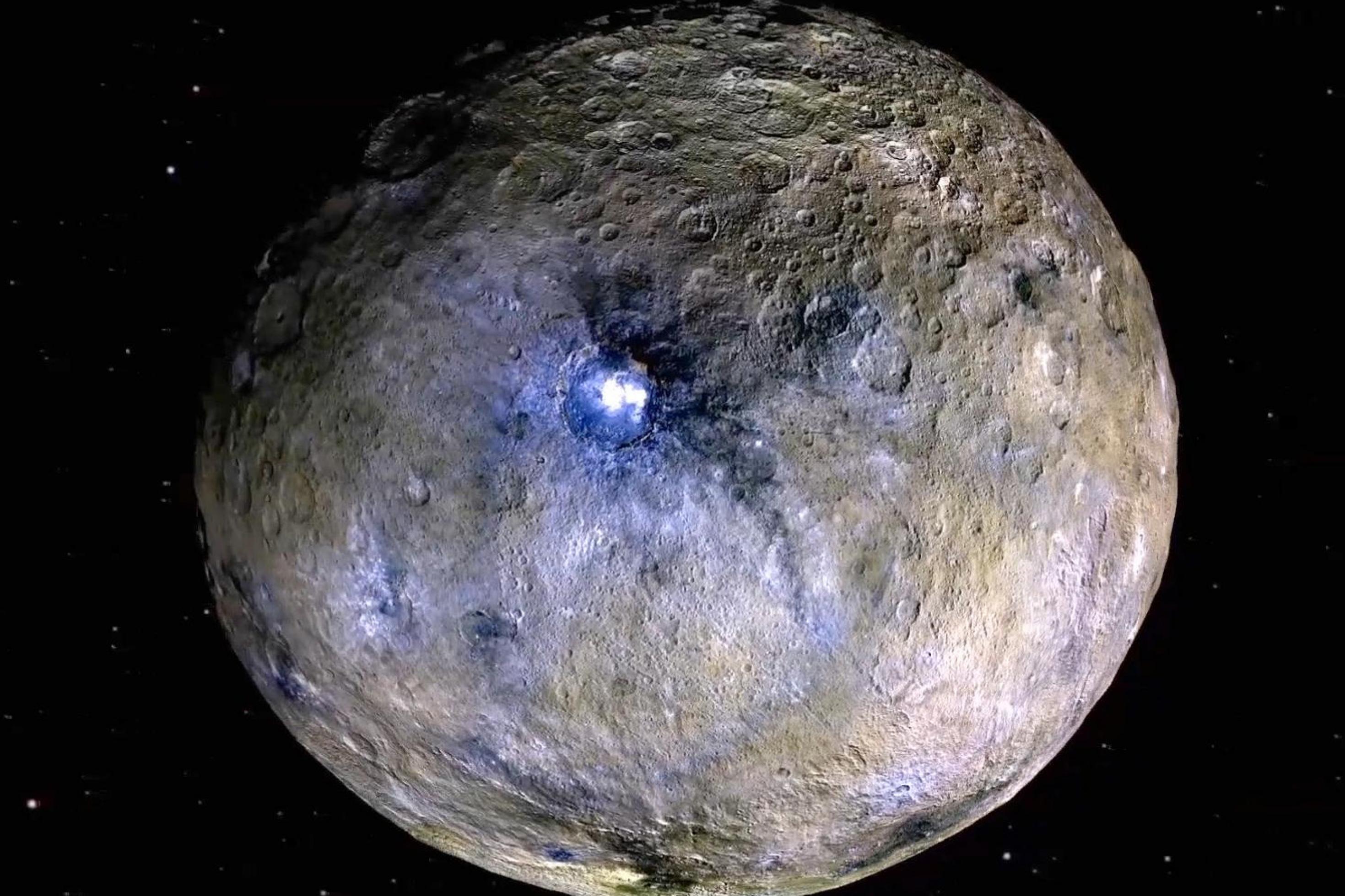 Der fast runde Planet in Gänze: braune und graue Farben und viele Krater, in der Mitte ein großer Krater mit gleißend hellen Flecken, der schwarz umrandet ist.