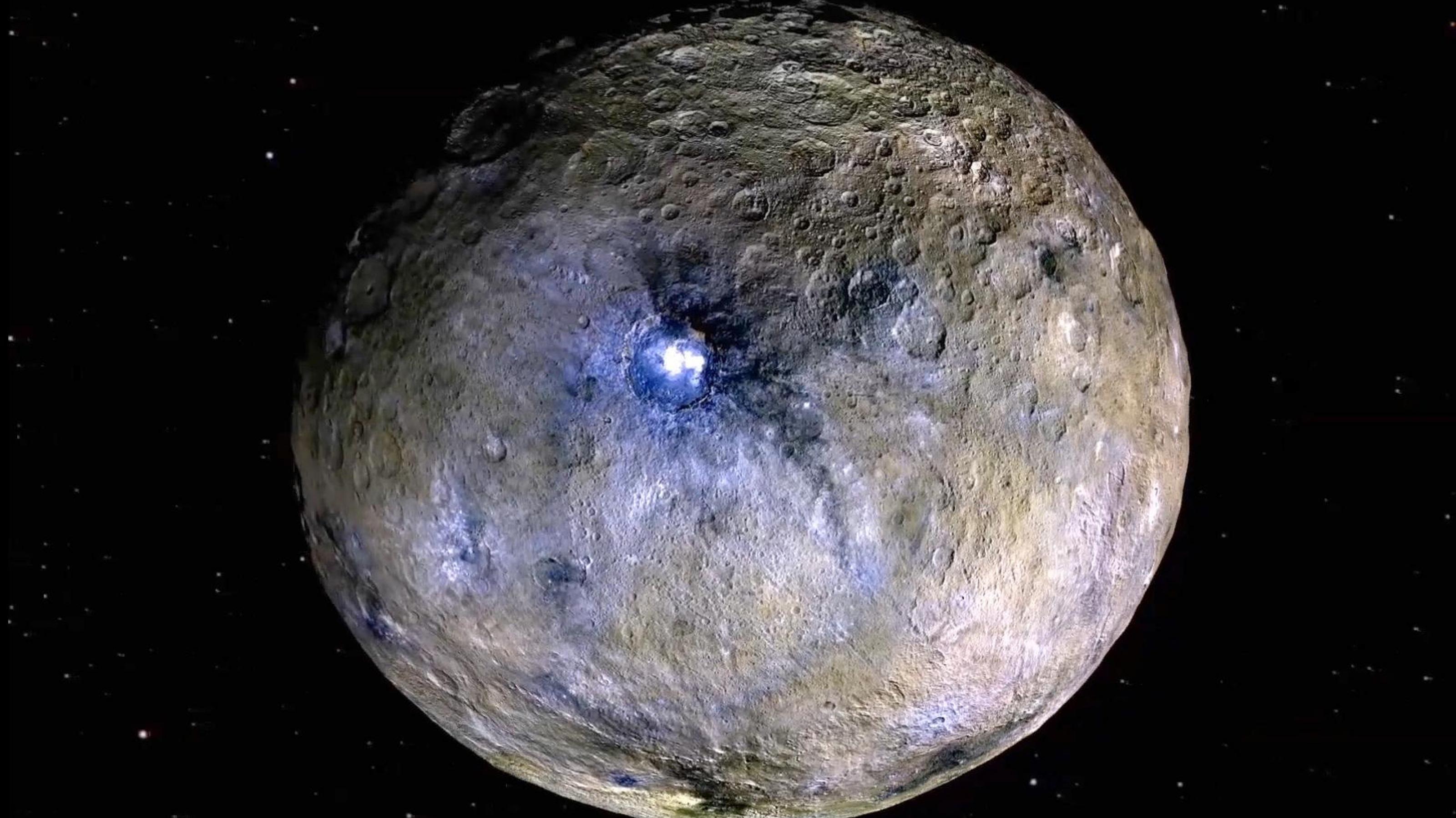 Der fast runde Planet in Gänze: braune und graue Farben und viele Krater, in der Mitte ein großer Krater mit gleißend hellen Flecken, der schwarz umrandet ist.