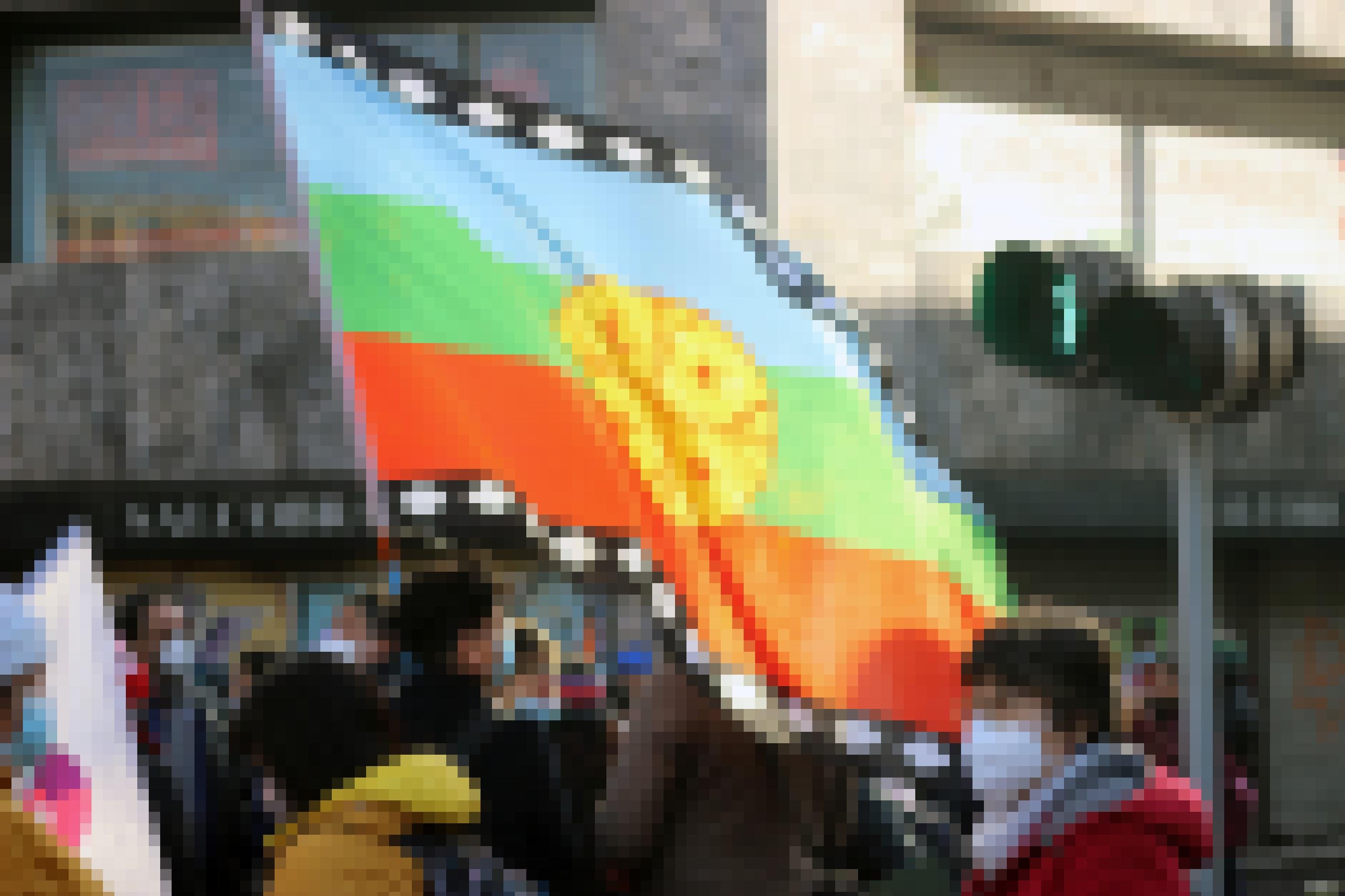 Auf der Straße stehen neben einer Ampel Menschen mit Mundschutz und halten eine bunte Fahne.