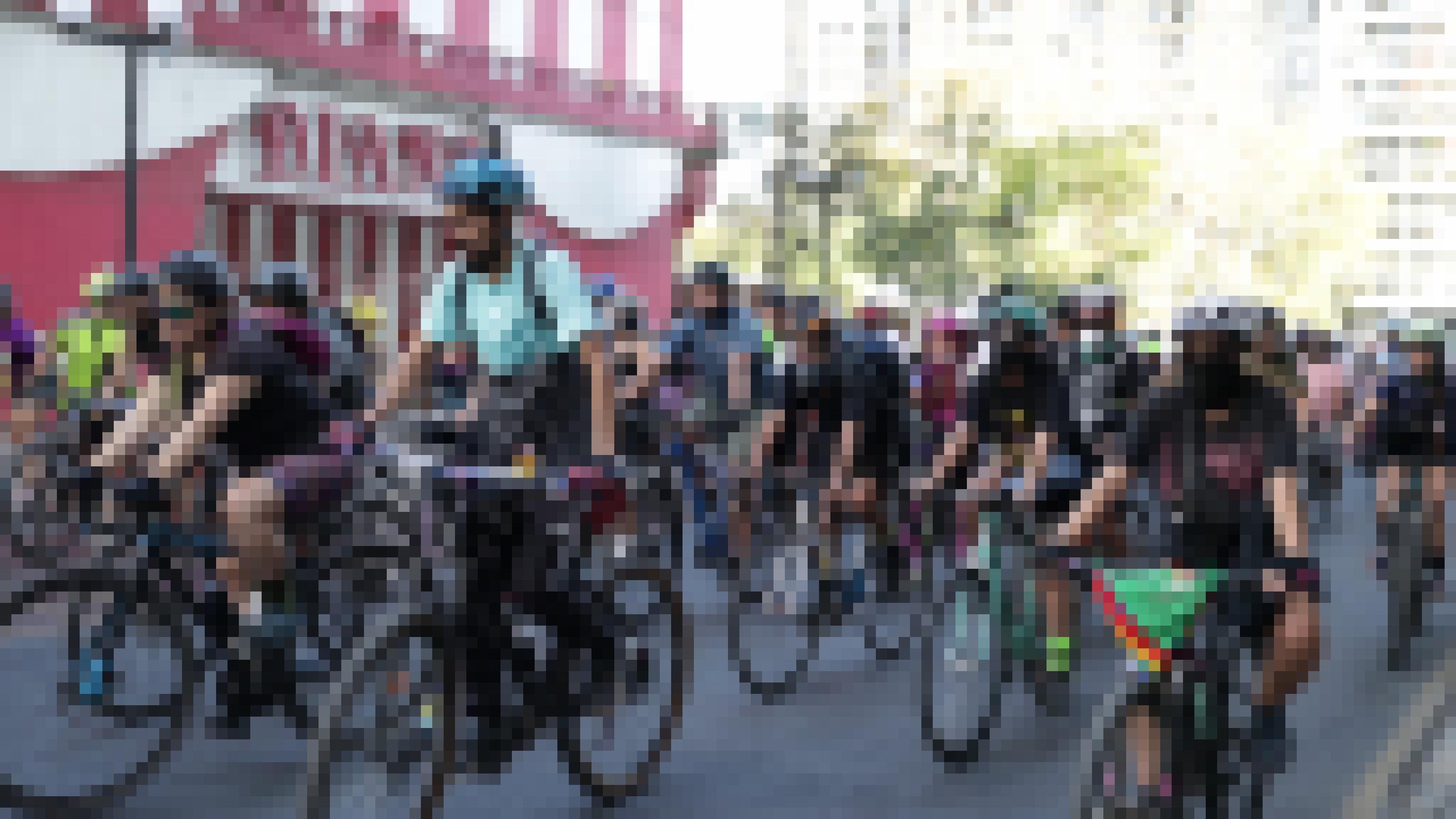 Eine Gruppe von Menschen, die Fahrrad fahren. Alle mit Helm, viele mit Sonnenbrille, manche Mundschutz. Sie sind für warmes Wetter gekleidet.