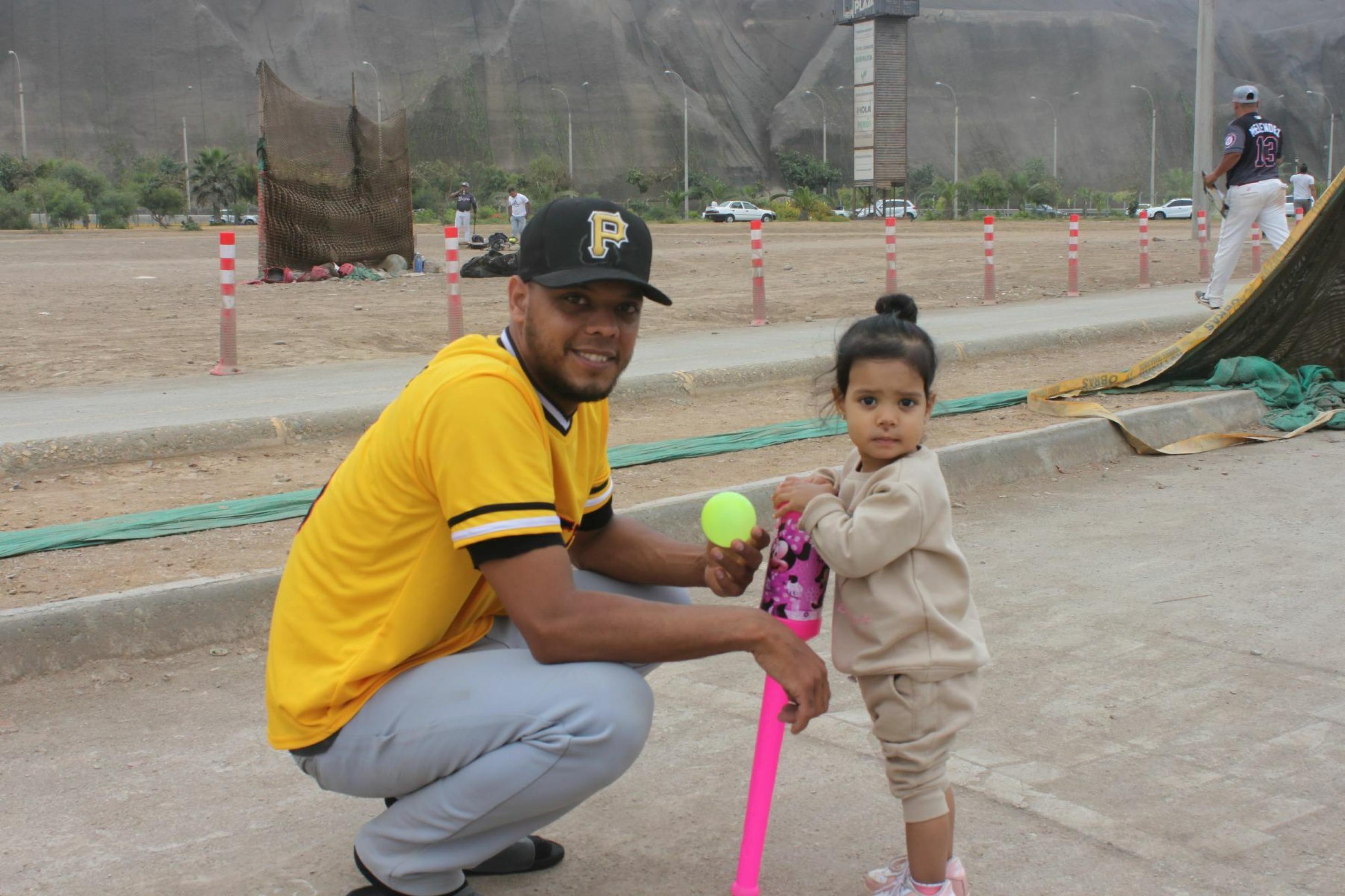 Links kniet ein junger Mann, braune Haut und Bart, mit Baseball-Mütze, vor ihm ein zweijähriges Mädchen, das in die Kamera lacht. Das Mädchen hält einen Mini-Plastik-Baseballschläger in der Hand. Der Mann zeigt ihr den gelben Ball.