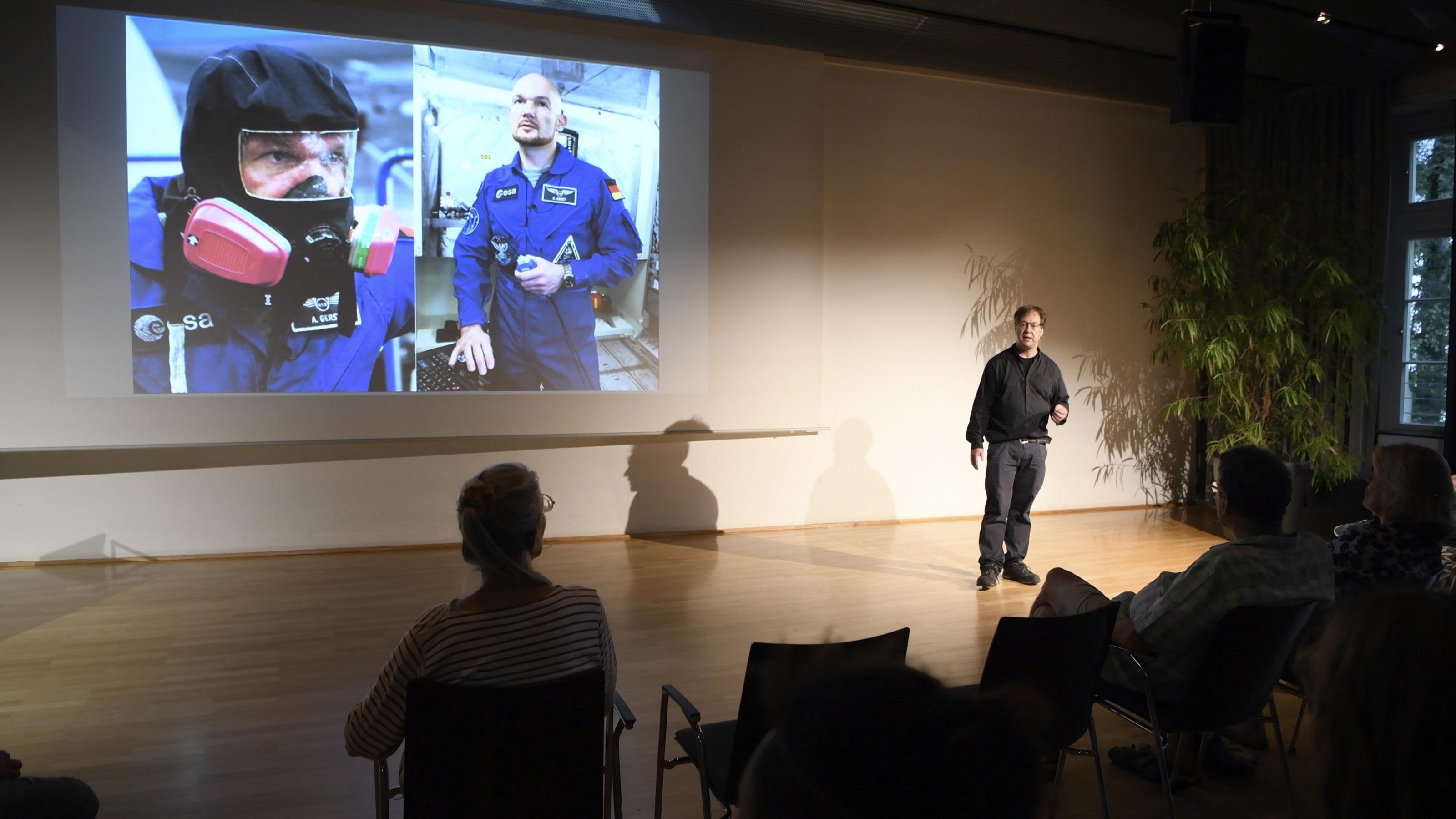 Christoph Otto auf einer Bühne, der Projektor zeigt zwei Bilder von Astronaut Alexander Gerst, eines davon mit Tauchermaske