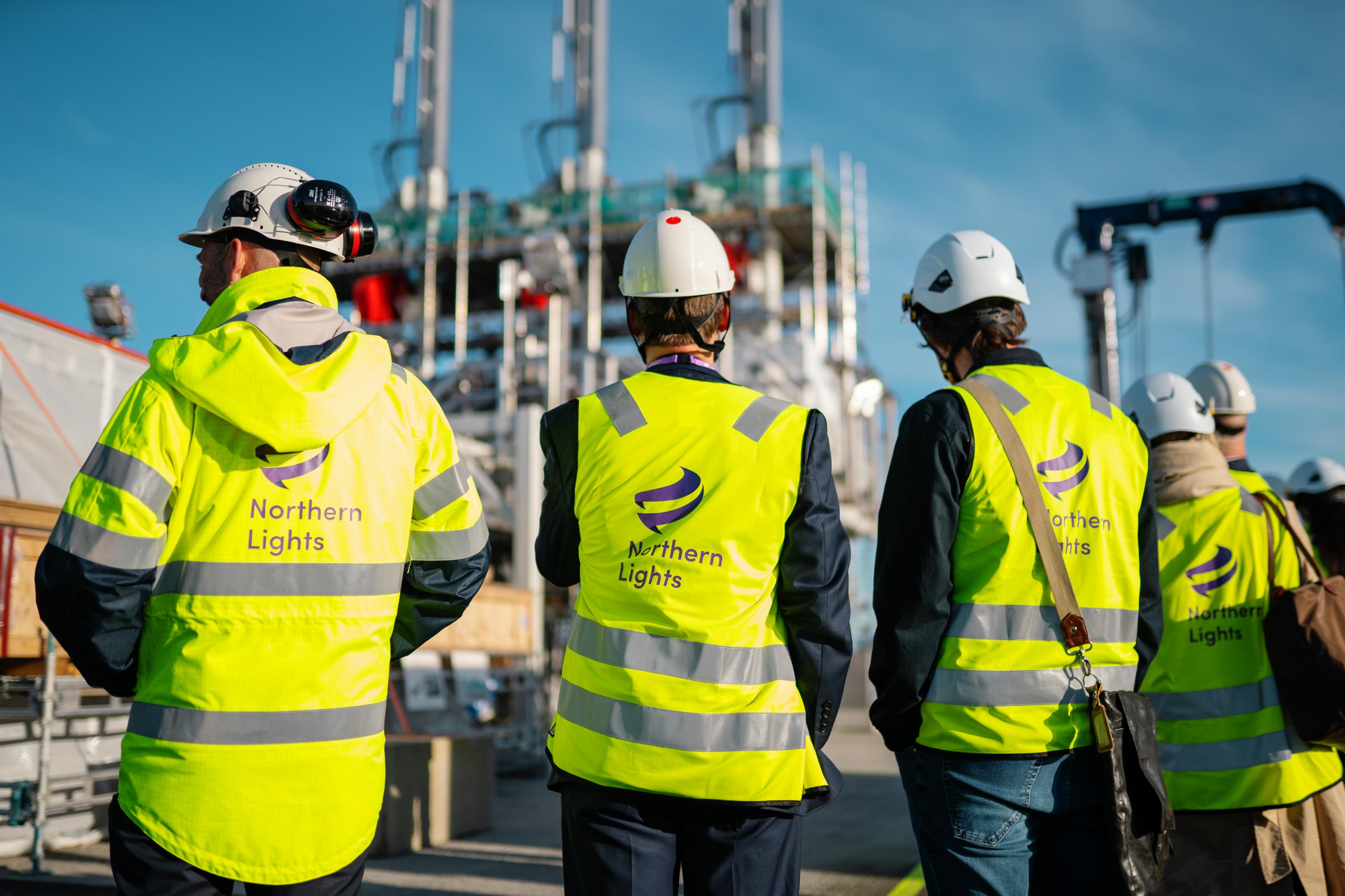 Vier Personen mit gelben Westen stehen mit dem Rücken zur Kamera vor aufrecht stehenden Stahltanks und anderen technischen Anlagen.