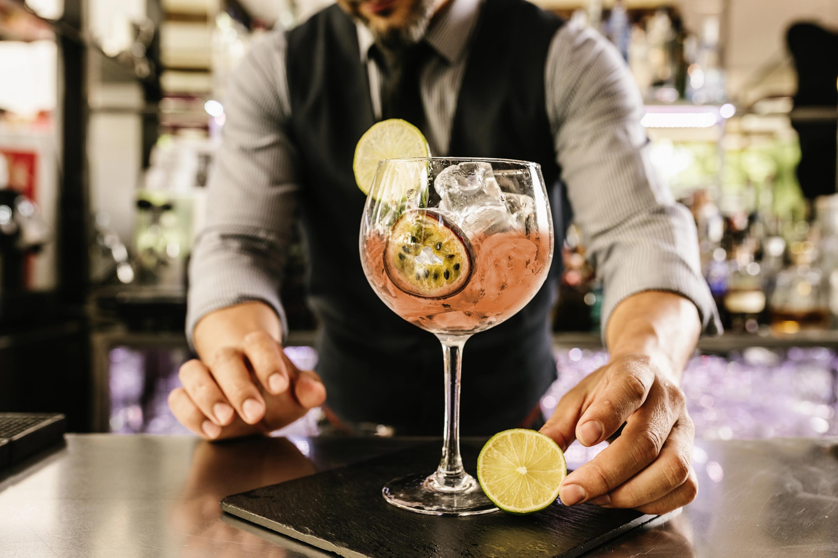 Halbnahe: Bartender in Unschärfe, alkoholfreier Aperitif scharf im Vordergrund, darin Eiswürfel und eine Scheibe Passionsfrucht.