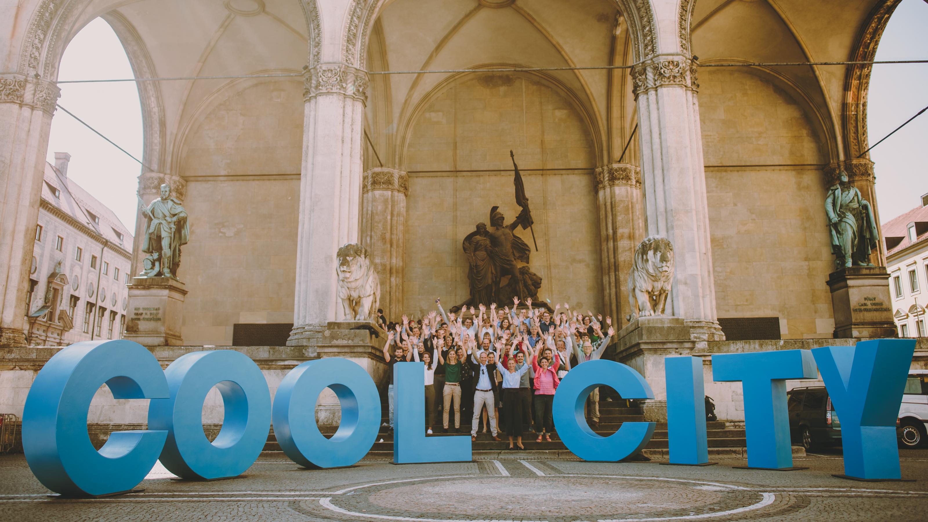 Eine Menschengruppe jubelt. Vor ihr steht Buchstaben aus Plastik, die das Logo der Stromsparprämie Cool City München formen