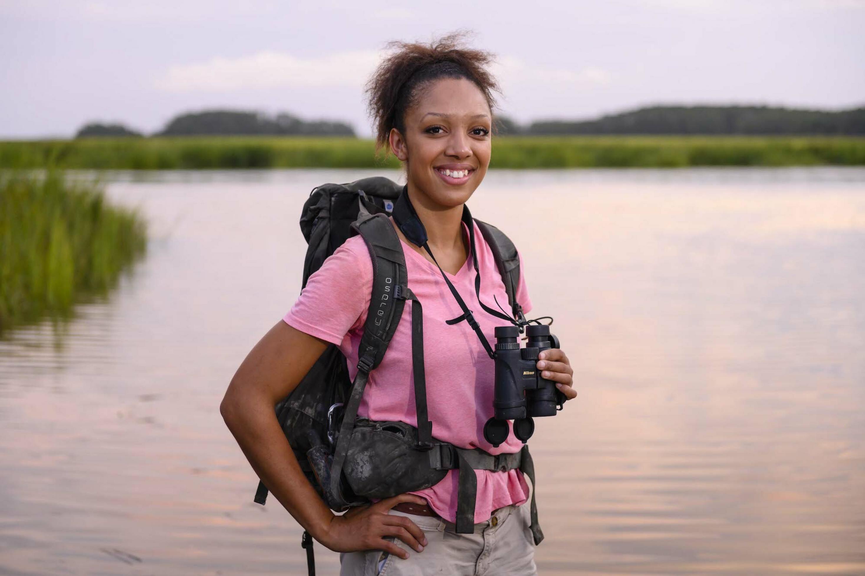 Zu sehen ist Corina Newsome mit einem Fernglas um den Hals in der Natur, vor einem Gewässer.