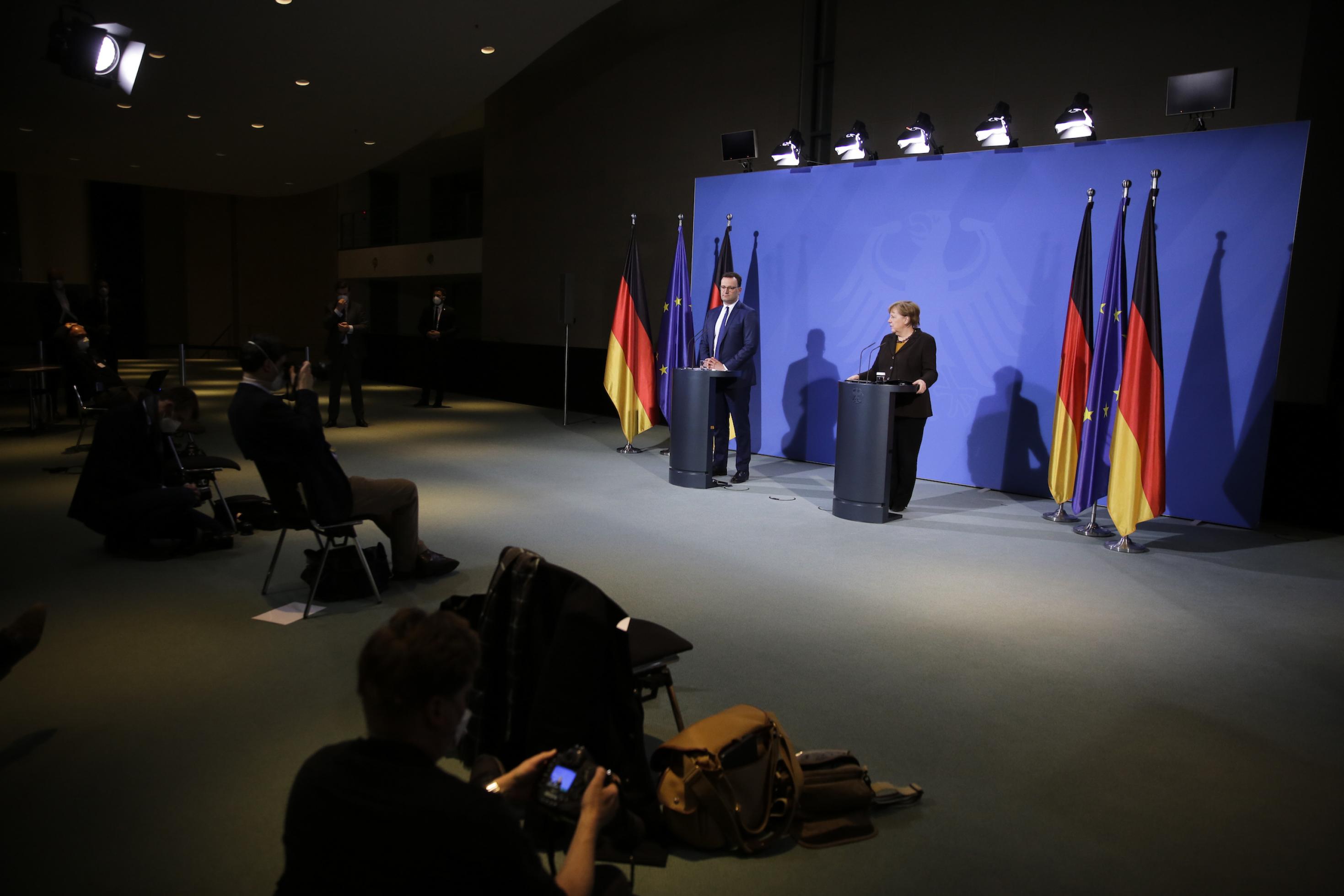 Merkel und Spahn stehen an Redepulten vor blauem Hintergrund, vor ihnen ein leerer Saal mit wenigen Journalistïnnen.