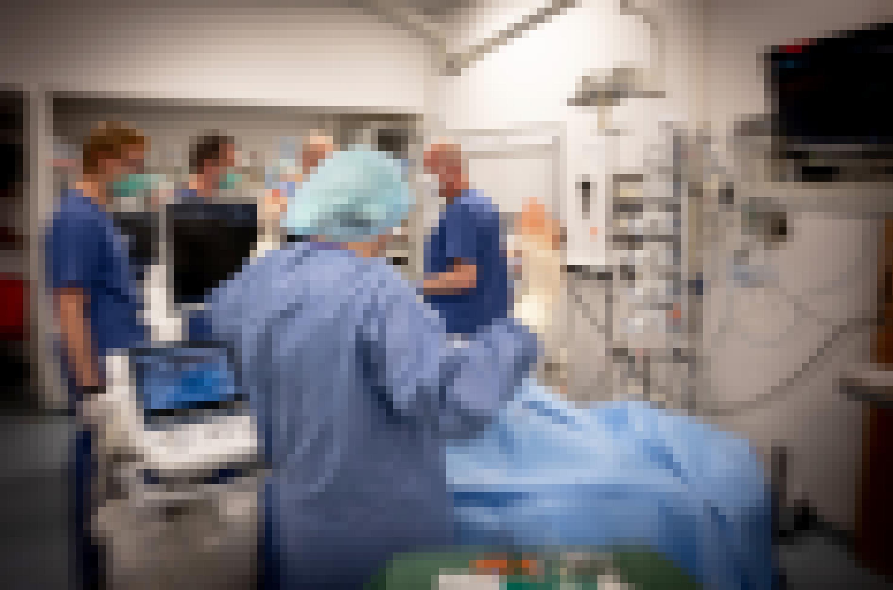 Ärztïnnen und Pflegerïnnen in Schutzkleidung auf einer Intensivstation zwischen medizinischen Geräten und einem Patienten in blauem Laken.