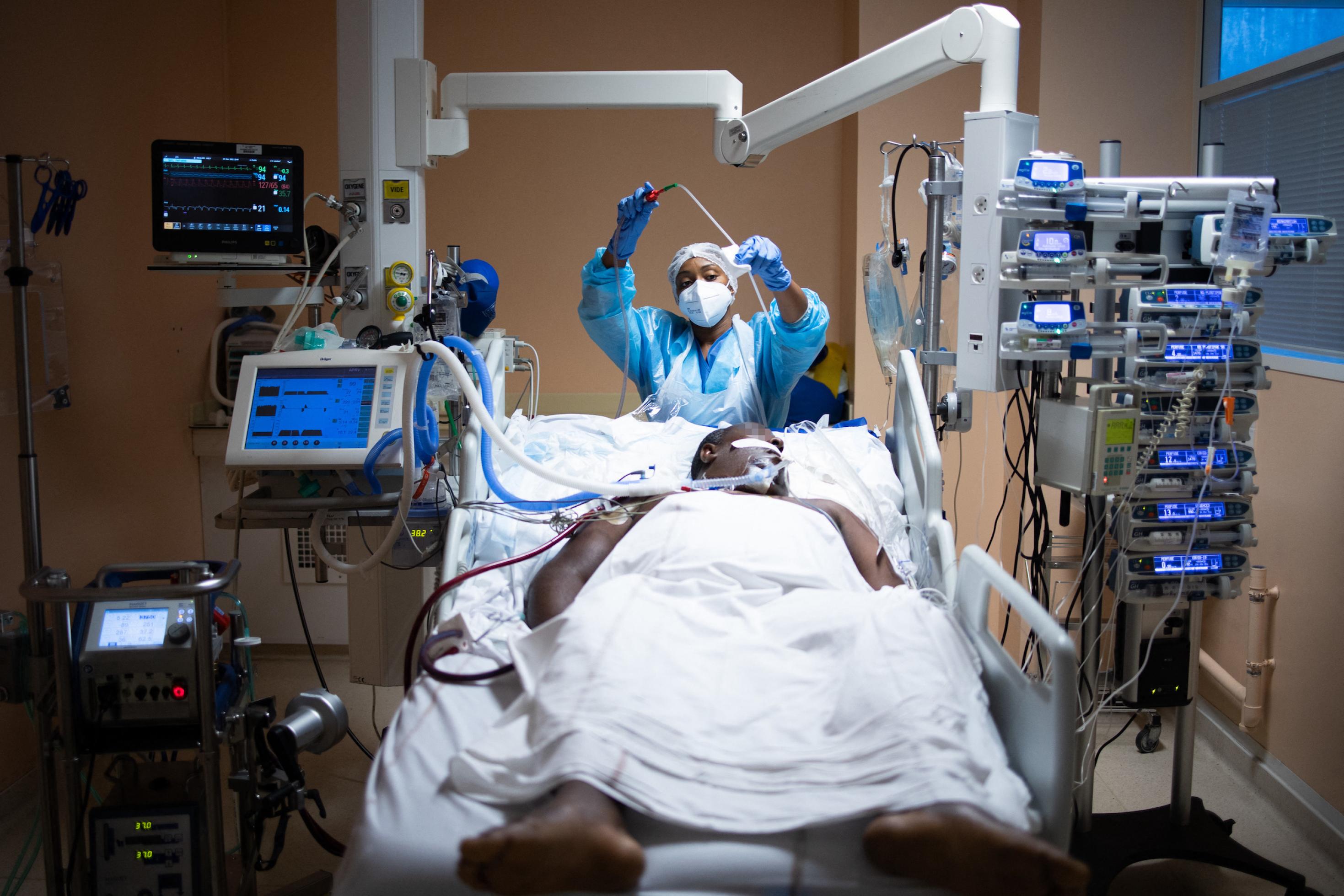 Ein Mann liegt in einem Intensivbett umgeben von medizinischen Geräten, eine medizinische Fachkraft steht am Bett und kümmert sich um ihn.