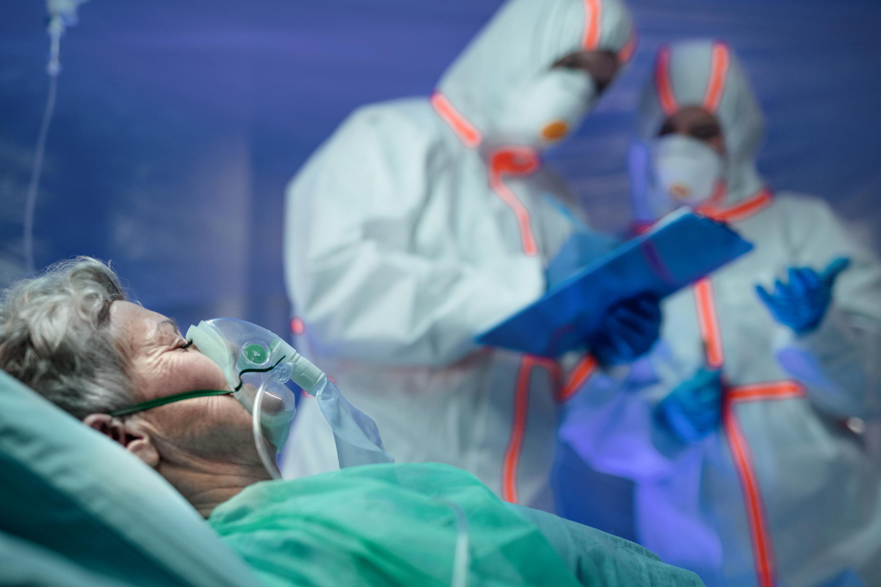 Eine ältere Frau liegt mit einer Atemmaske in einem Krankenhausbett. Im Hintergrund sieht man zwei Menschen, das medizinische Personal, im Schutzanzug.