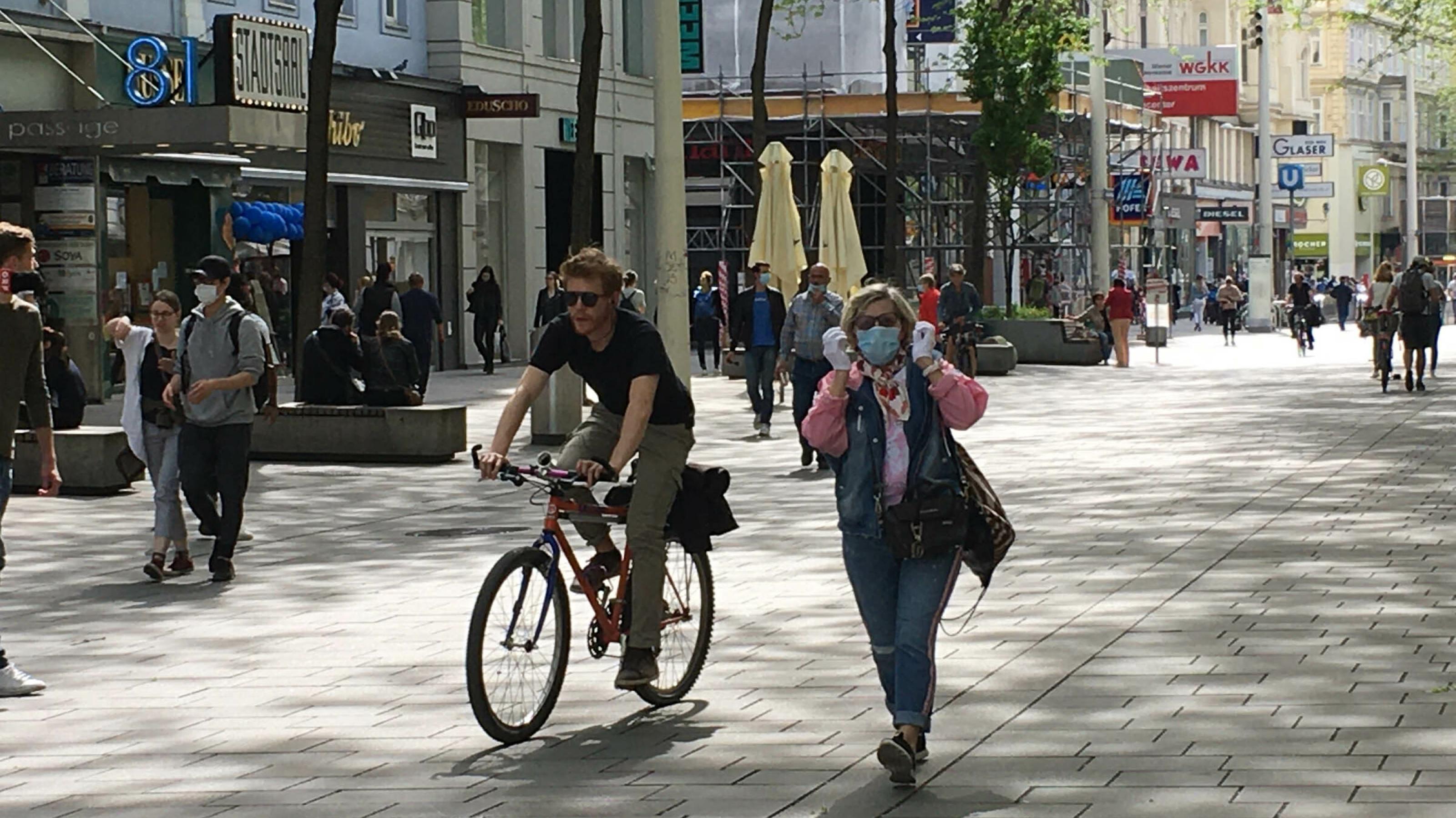 Einkaufsstraße mit Fußgängern und Radfahrern, eine Frau trägt Mundschutz.