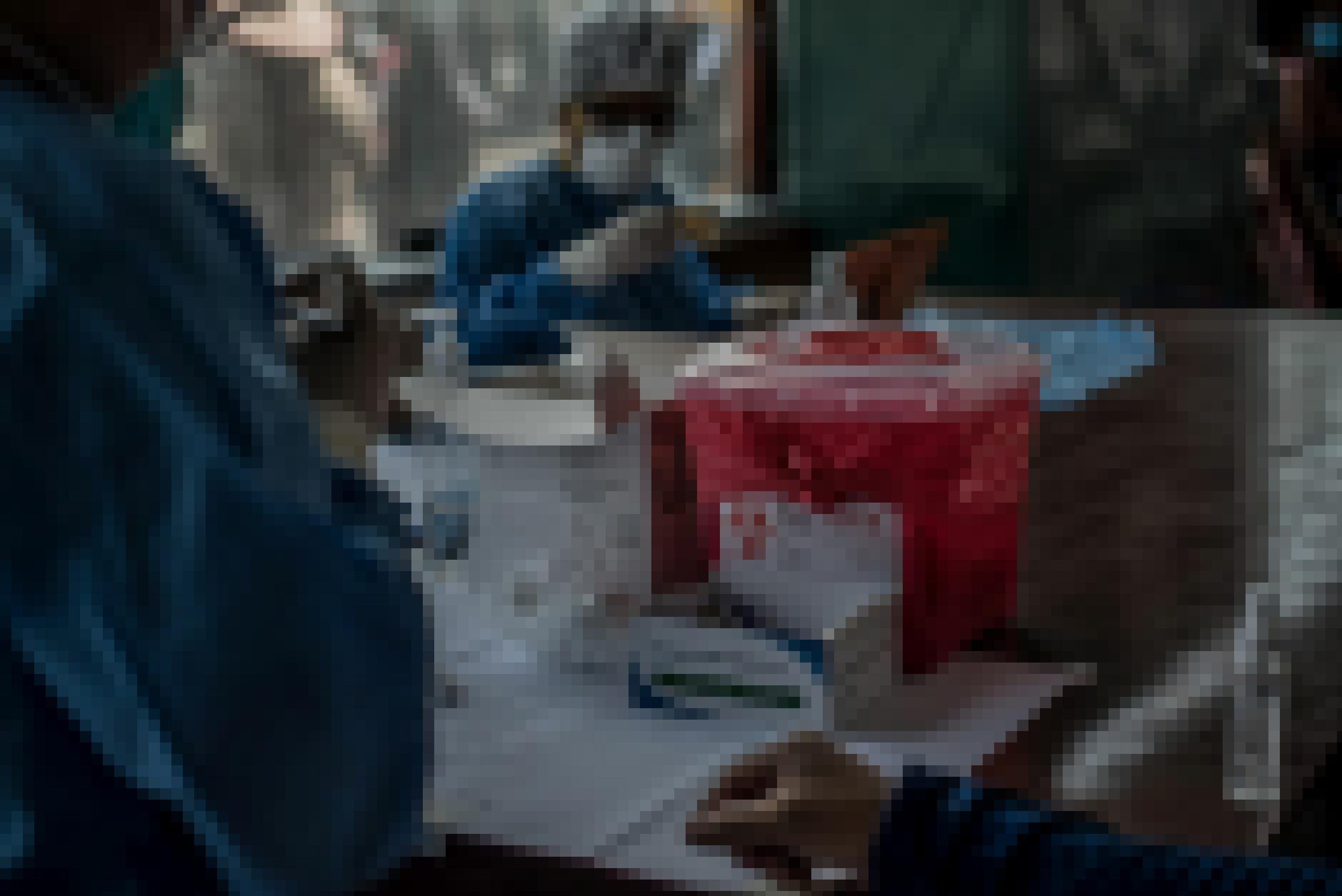 Im Hintergrund steht ein Mann in blauem Hygienekittel und Mundschutz, der eine Spritze aufzieht. Im Vordergrund steht ein kleiner roter Plastikeimer für gebrauchte Spritzen.