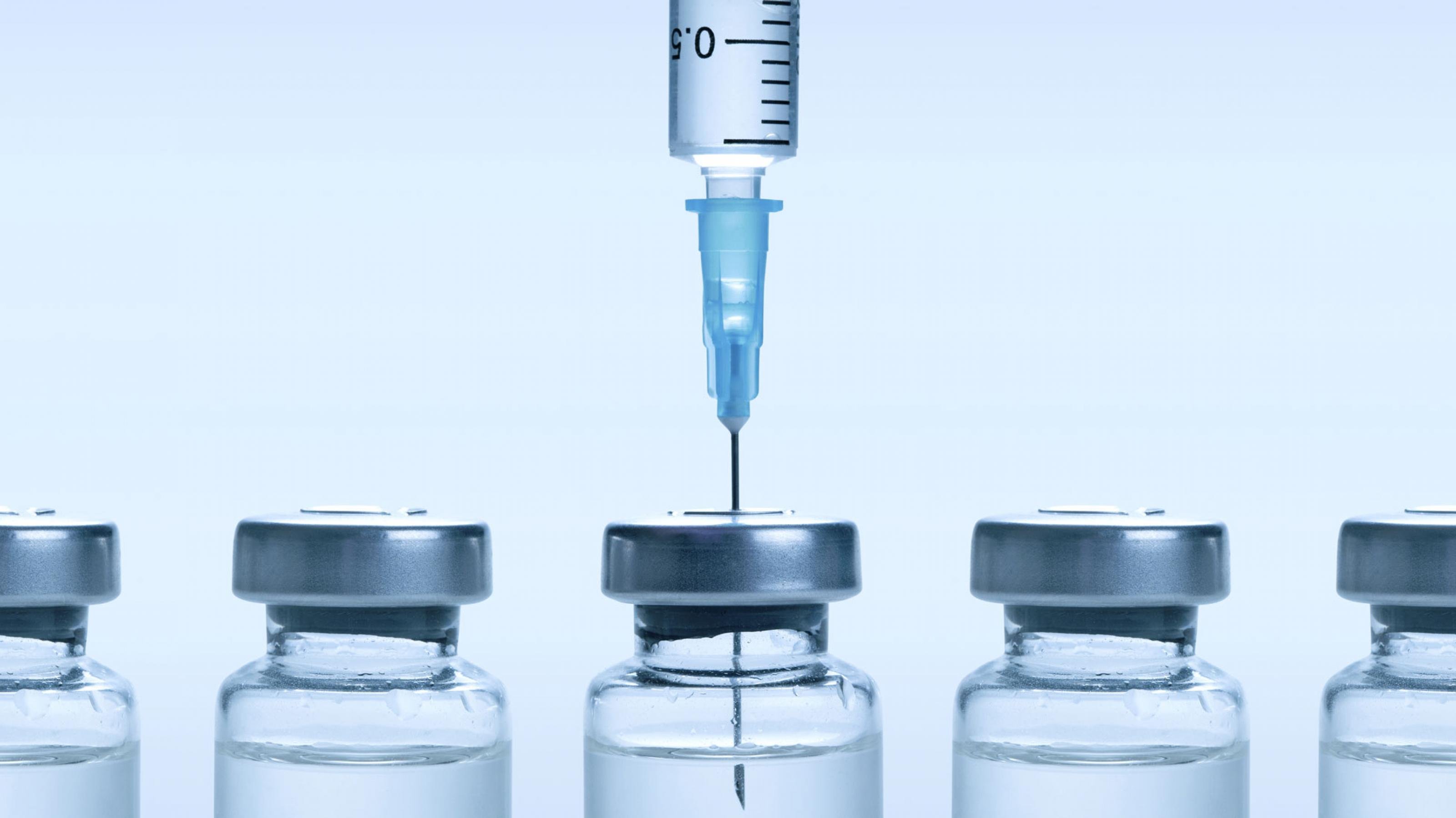 Das Bild zeigt fünf Impfstoff-Fläschen, aus dem mittleren wird der Impfstoff gerade mit Hilfe einer Spritze aufgezogen.